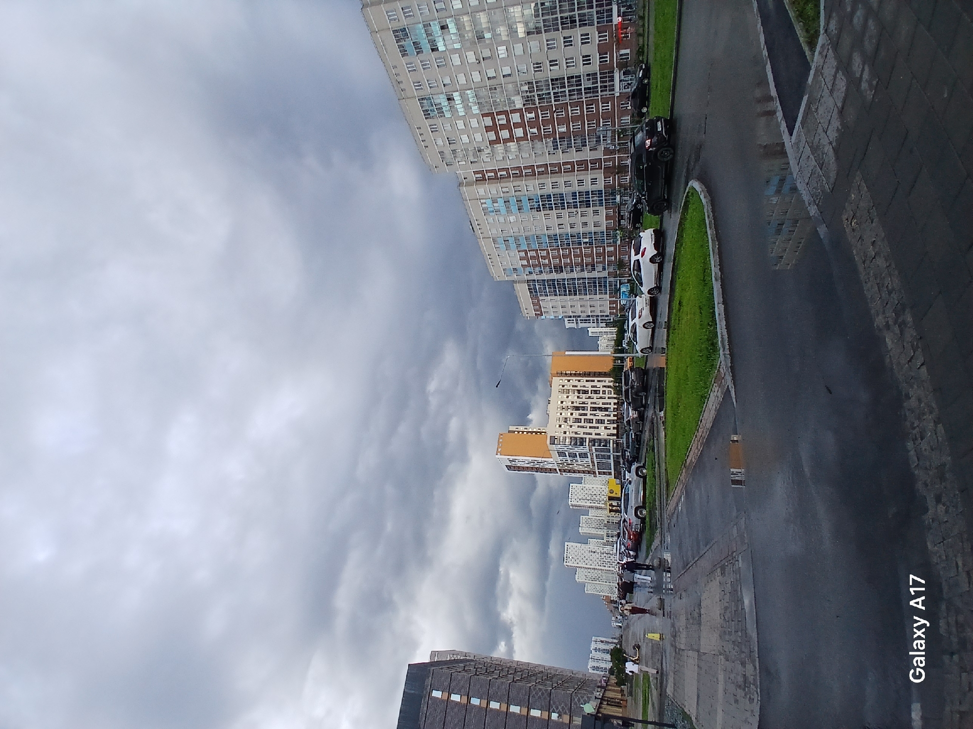 The image displays an urban residential street scene in Yekaterinburg, Russia, under a heavily overcast sky. The overall atmosphere is dim and damp, with wet asphalt and paved sidewalks reflecting the light, suggesting recent rain or high humidity.
Numerous multi-story residential buildings dominate the landscape, lining both sides of the street. These modern apartment blocks feature varied facades, with some showcasing light grey and white panels, while others incorporate brown and orange elements. Windows on the buildings reflect the cloudy sky.
A paved road stretches through the scene, flanked by narrow strips of green grass. A variety of cars, including black and white vehicles, are parked along the curbs. A wide, wet sidewalk runs along the right side of the image, where several people are seen walking. One person in a white top and dark pants is clearly visible. Reflections of the buildings are discernible in the puddles on the road and pavement.
The time of day is daylight, but the dense, dark grey clouds cover the entire sky, giving the scene a muted, pre-storm or post-rain appearance. The visible text "Galaxy A17" is present in the bottom right corner of the original image, indicating the camera used.