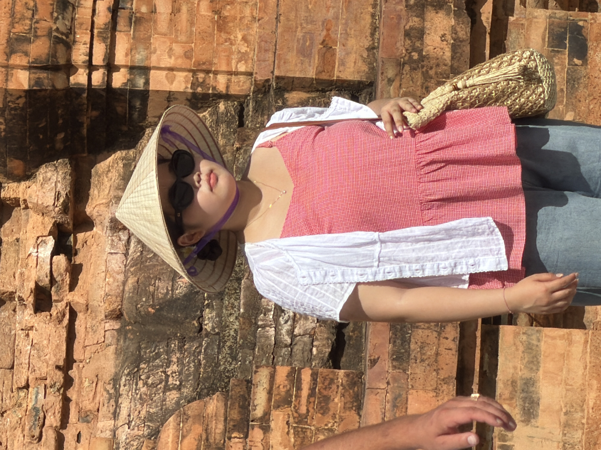 A young woman stands in front of a weathered brick wall in what appears to be the Po Nagar Cham Towers in Nha Trang, Vietnam. She wears a traditional conical hat adorned with purple ribbon, dark sunglasses, a white button-up shirt over a pink and white gingham print peplum top, and light blue jeans. She holds a woven straw bag with tassels in her right hand. A man's arm, wearing a ring, is visible on the left side of the frame, suggesting they are together. The brickwork of the wall shows signs of age and possible water damage, with darker stains and uneven surfaces. The lighting indicates a bright, sunny day.