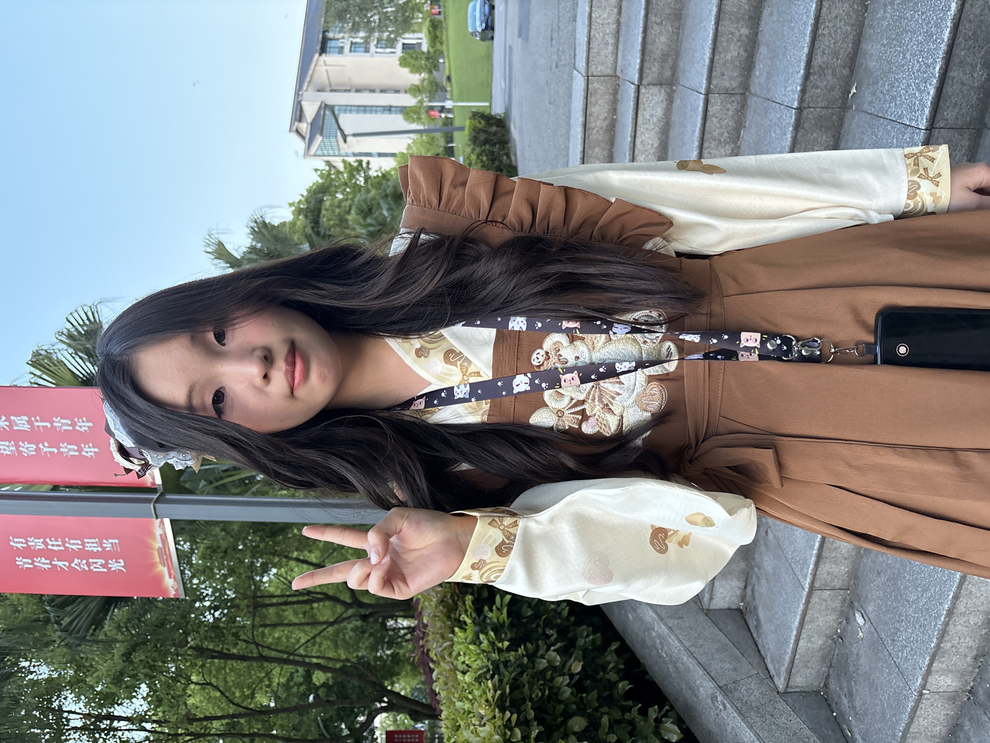 A young woman stands in Jiangjin, China. She is wearing a brown and cream-colored dress with ruffled detailing and a lanyard featuring cartoon animals. She is holding up a peace sign. The background includes trees, buildings, and stone steps. Red banners with Chinese text are also visible.