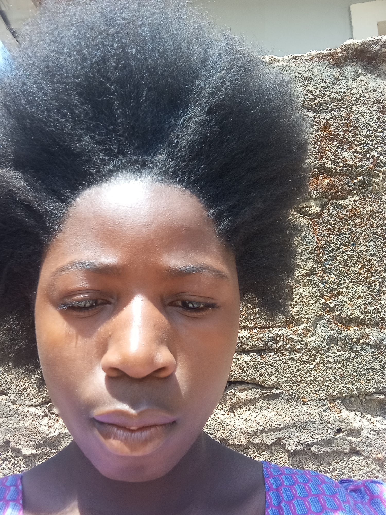 A close-up selfie of a dark-skinned individual with natural, afro-textured hair stands against a textured wall. The subject's face is prominent, showing a slight downward gaze, and their lips are lightly pursed. The rough texture of the wall contrasts with the smooth skin of the face. The environment appears to be outdoors, with natural light casting shadows on the face and wall. Based on the prompt specifying location in Jos, Nigeria, the wall is likely a common building material and style in the region.