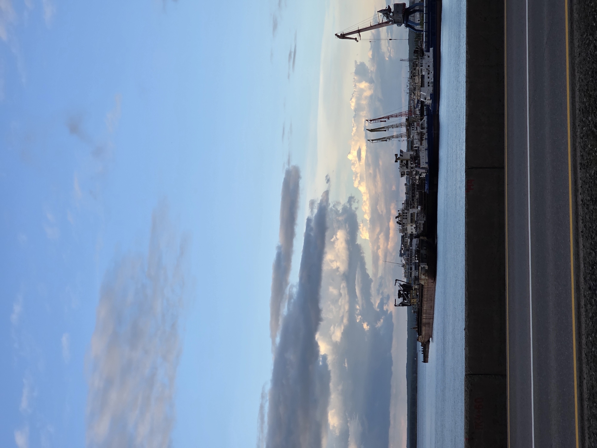 This image depicts a port scene in Chaikovsky, Russia, during what appears to be late afternoon or early evening, with a dramatic sky filled with clouds illuminated by the setting sun. The primary subjects are a collection of ships and cranes lined up along the waterfront, suggesting ongoing port operations. Several large cranes, including a prominent red one at the top of the frame, tower over the docked vessels. The water in the foreground is calm, reflecting the soft light of the sky.
To the right of the water is a concrete barrier and a road with distinct white and yellow lane markings. The road is dark, indicating it might be asphalt. The overall atmosphere is one of industrial activity juxtaposed with the beauty of the natural sky. The clouds exhibit a range of colors from soft blues and grays to warm oranges and yellows, hinting at a sunset.
There is some visible text on the concrete barrier in the lower right quadrant of the image, which appears to be markings or graffiti. The text is difficult to read clearly but seems to include numbers and possibly Cyrillic characters. No people are explicitly visible in the image, and the focus is on the infrastructure of the port and the surrounding environment. The scene conveys a sense of scale and the operational nature of the port.