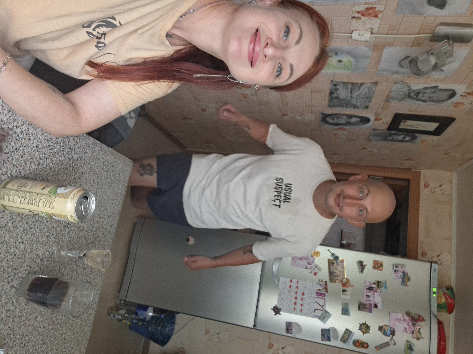 The image captures a domestic scene, likely an apartment kitchen, where a woman with red hair is taking a selfie in the foreground. She is smiling and wearing a light-colored t-shirt with a dark design. Behind her, a bald man in a white t-shirt that reads "USUAL SUSPECT" and dark shorts is visible. He appears to be reacting with surprise or amusement. The background reveals a wall adorned with drawings and photographs, a refrigerator covered in magnets and papers, and a doorway. On a speckled countertop in the foreground, a can of beverage, a small liqueur glass, and a glass filled with a dark liquid are placed. The overall impression is of a casual, possibly informal gathering or moment. The location of Myadzyel, Belarus is not discernable from the visual elements present in the image.