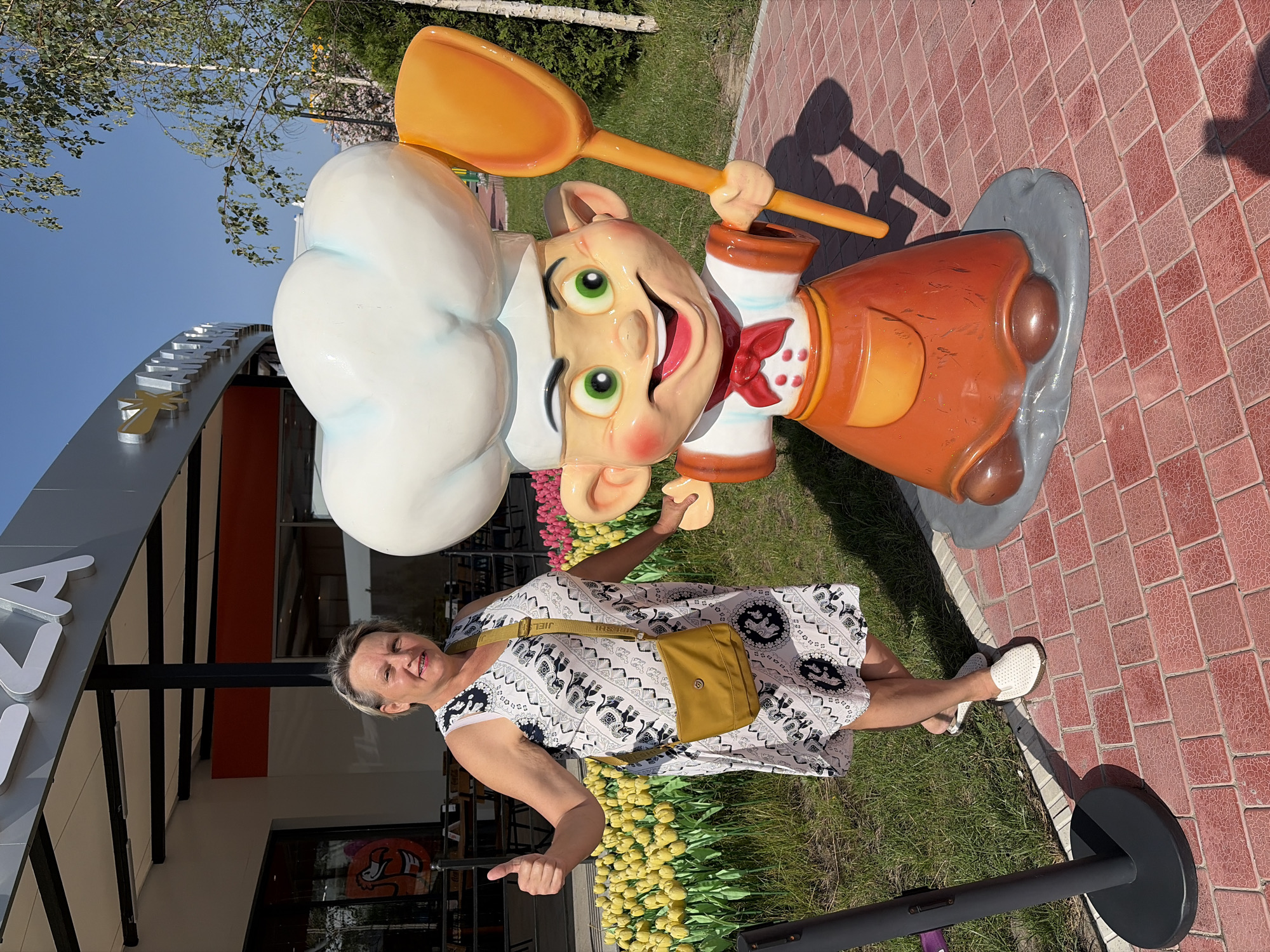 A middle-aged Caucasian woman with blonde hair smiles and gives a thumbs-up while posing for a photo next to a cartoonish chef statue. She is wearing a sleeveless white dress with a black and gray elephant print, a yellow crossbody bag, and white perforated loafers. The statue, which is much larger than the woman, depicts a character with a large white chef's hat, green eyes, a red bow tie, and an orange apron, holding a large orange spatula. They are standing on a red brick pathway in front of a building with "PIZZA" visible on a sign. In the background, a large patch of yellow tulips and other flowers can be seen, along with trees and a clear blue sky. The scene appears to be outdoors in a park or entertainment area.