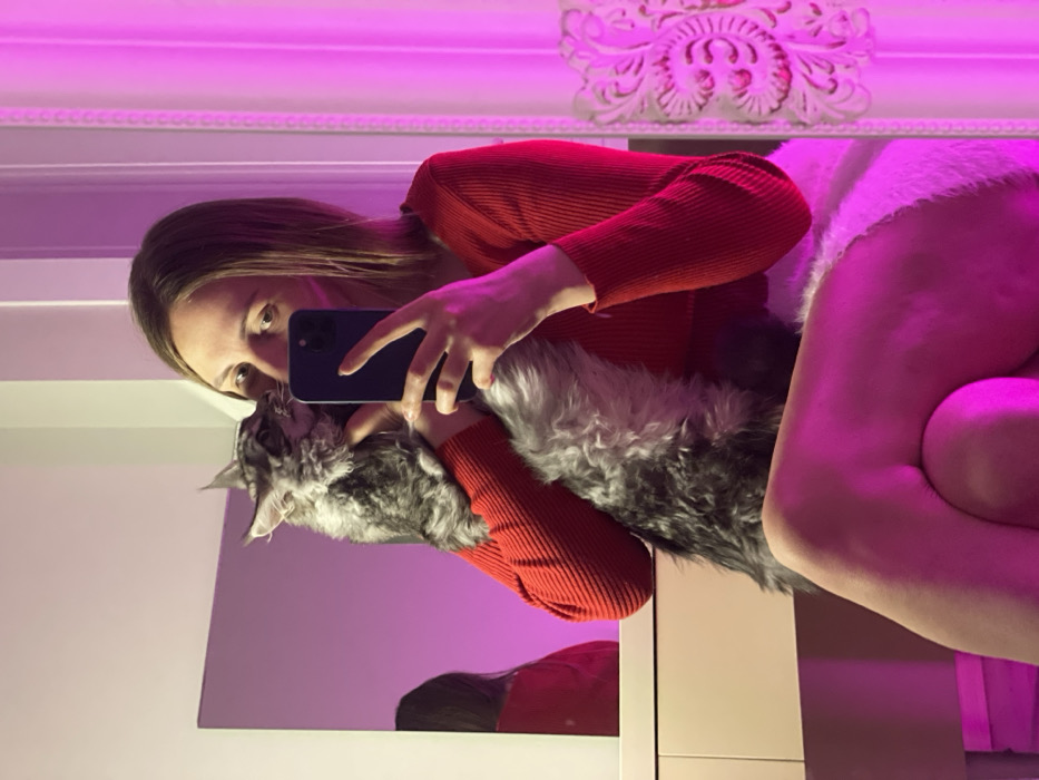 A woman in a red sweater and white shorts takes a selfie in a mirror. She holds a large gray Maine Coon cat close to her face, seemingly kissing it. The image is bathed in pink light, illuminating a decorative wall element above. The woman's reflection is visible in the mirror, alongside the cat. She stands in front of white furniture.