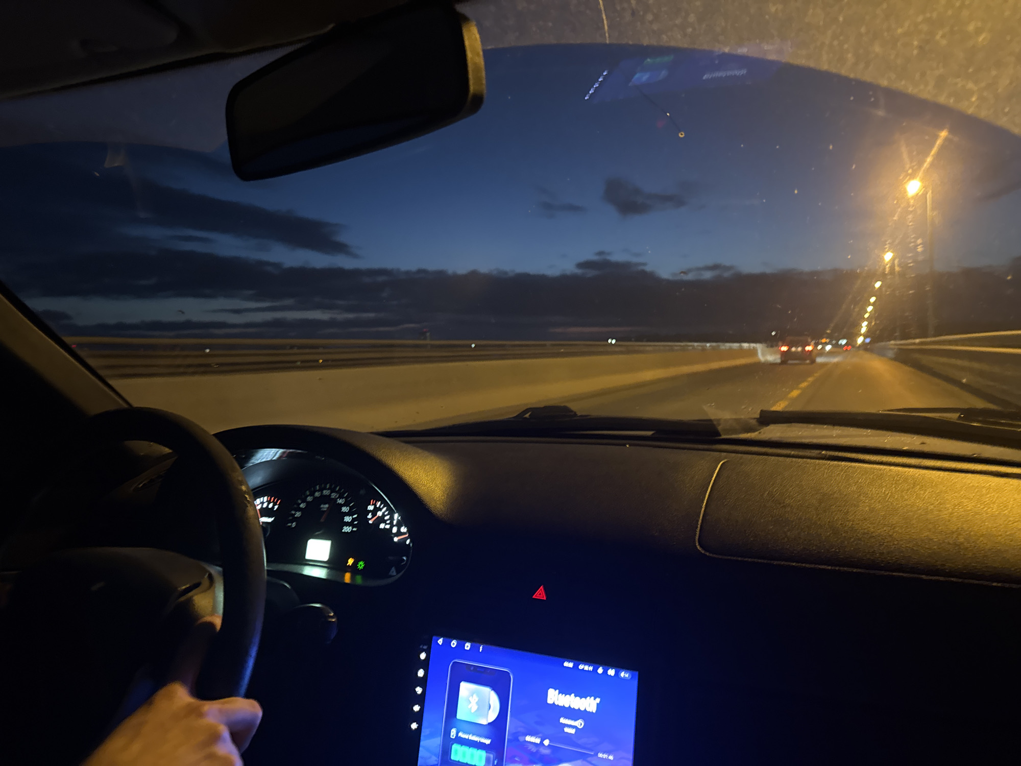 The image depicts the interior of a car at dusk, with a view through the windshield onto a highway. The driver's hand is visible on the steering wheel on the left. The dashboard features a speedometer, a touchscreen infotainment system displaying a Bluetooth connection, and a hazard light indicator. Streetlights line the highway, illuminating the road ahead, where other vehicles are visible in the distance. The sky is a deep blue, with scattered clouds. The scene suggests the car is in motion on a road that appears to be an elevated highway, possibly a bridge, due to the presence of guardrails and the clear expanse of sky. The overall impression is that of a nighttime drive on a well-lit road outside of a city.