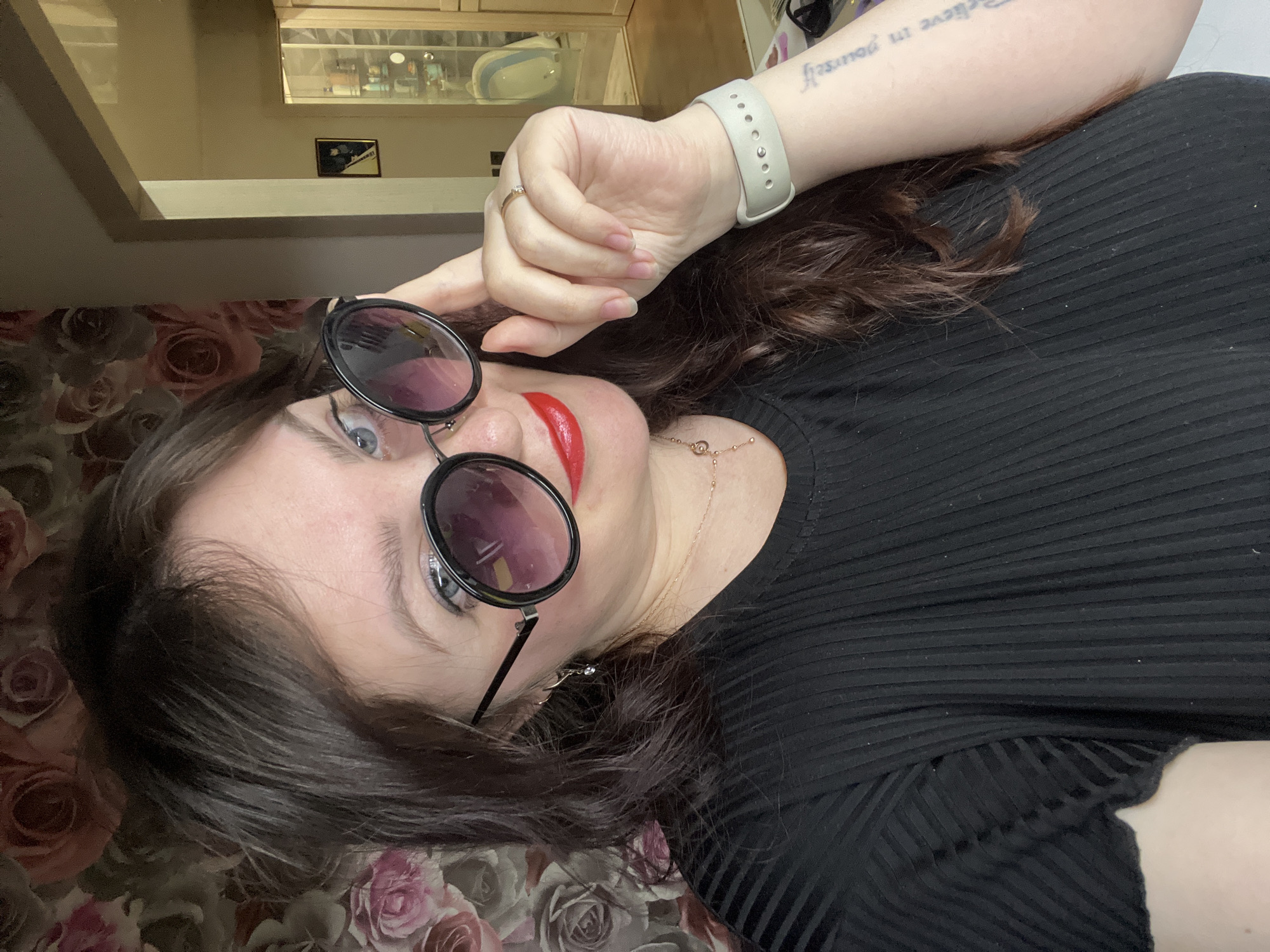 An adult woman is depicted in a medium close-up indoors in Noviy, Russia. She has dark brown, wavy hair and fair skin, with blue eyes partially visible above large, round-framed black sunglasses featuring dark gradient lenses. Her lips are covered in red lipstick. She wears a black, short-sleeved, ribbed top. Her left hand is raised, with the index finger touching the rim of her left sunglass lens. On her left ring finger, a gold-colored ring with a clear stone is present. A light grey silicone watch band is on her left wrist, and a tattoo with partial text "in yourself" is visible on her left forearm. A delicate gold-colored necklace with small pendants, including a circular one and a cross, is around her neck, and a small, clear-stone earring hangs from her left ear. The background on the left consists of wallpaper patterned with muted pink and brown roses. To the right, an interior doorway leads to another room with light-colored walls. Inside this room, a framed picture of a black cat hangs on the wall, and a tall, glass-fronted cabinet containing various items is visible, along with a partially obscured white object.