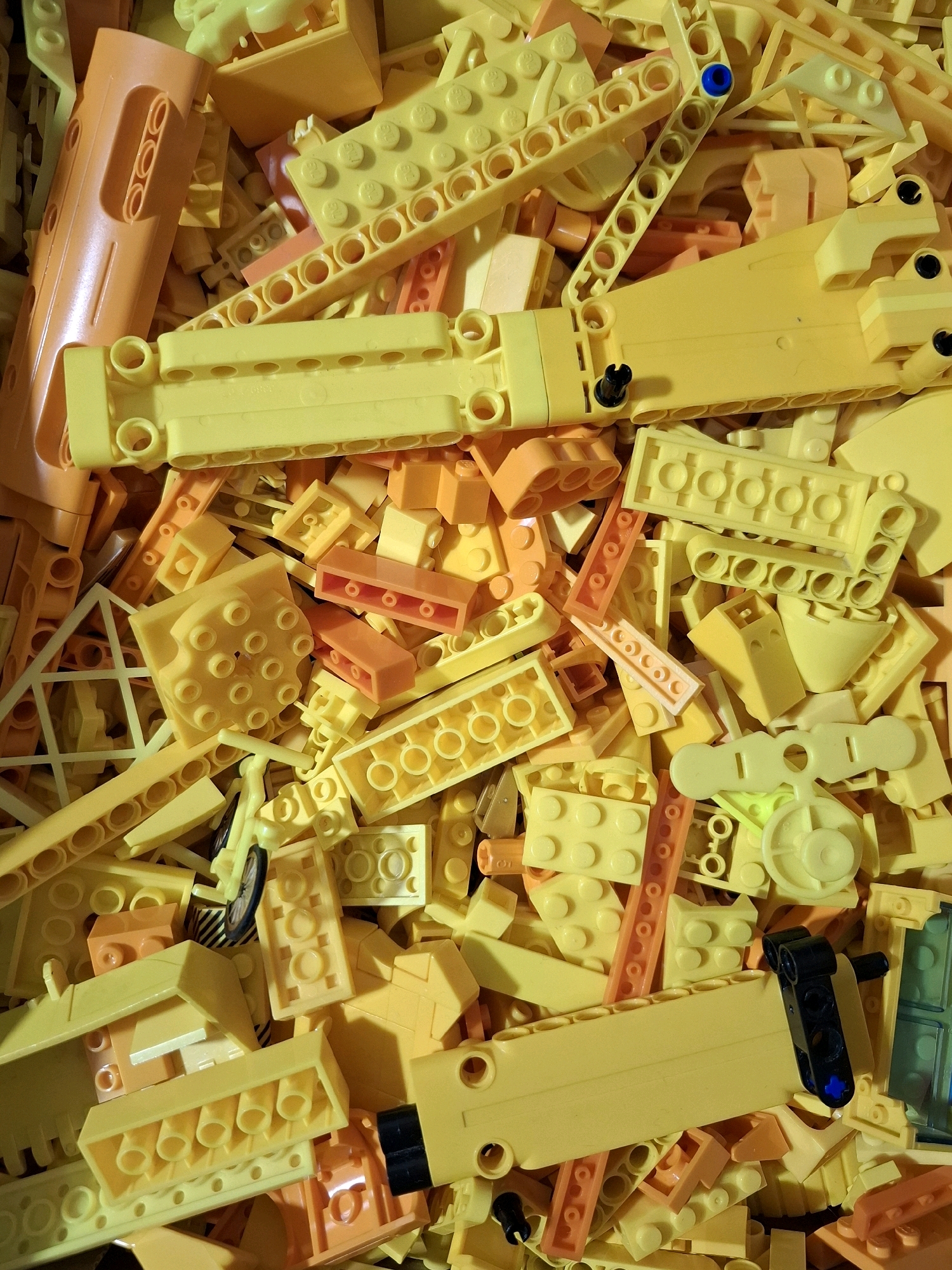 This image is a close-up, top-down view of a large pile of yellow and orange plastic building blocks. The blocks vary in size and shape, including long rectangular pieces with circular holes, smaller square and rectangular bricks, and some specialized pieces with various connectors and curved elements. Some blocks have visible circular stud patterns. There is a small black and blue piece attached to one of the yellow blocks in the lower right quadrant. The lighting creates some shadows and highlights, emphasizing the textured surfaces of the plastic. The scene is entirely composed of these toy blocks and does not depict any specific location, including the city of Tyumen, Russia. There are no people, animals, or any other objects present.
