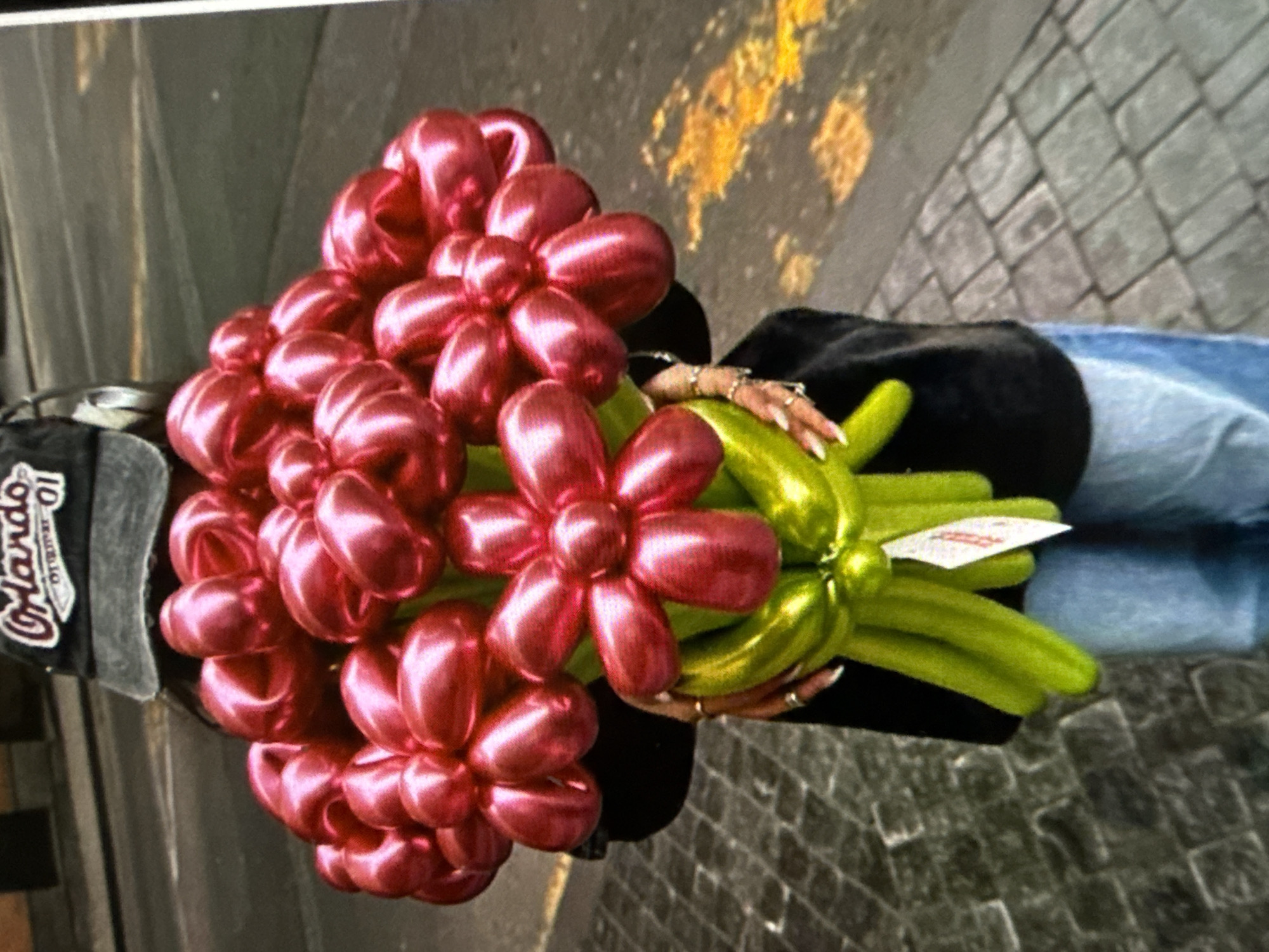 The image depicts a bouquet of pink flower-shaped balloons with green stems, held by a person wearing blue jeans, a black top, and an Orlando 01 baseball cap. The location appears to be an outdoor setting with a brick paved surface. The person's hands, adorned with rings, are visible holding the balloon bouquet.