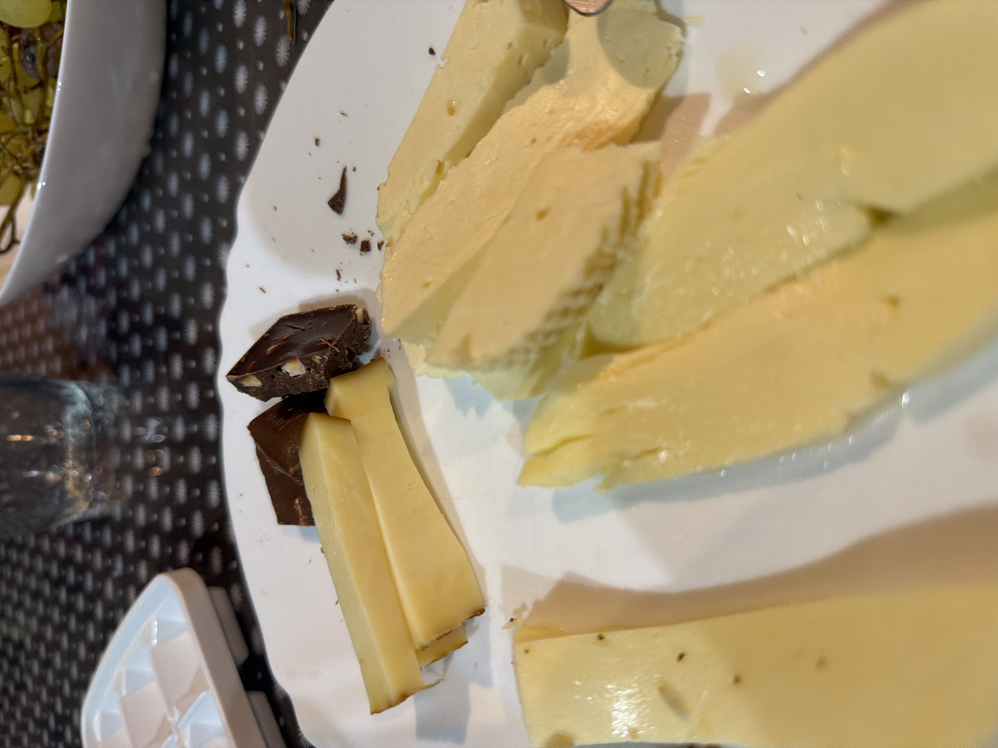 On a white plate, several slices of light yellow cheese are arranged. Two pieces of dark chocolate, one with visible nuts, are also on the plate. The plate rests on a dark patterned tablecloth. In the background, an ice cube tray is visible on the left, and a glass with a clear liquid is on the right. A bowl containing grapes is also partially visible in the upper right. The scene depicts food on a table, likely in a domestic setting. There is no indication of the city of Sochi, Russia.