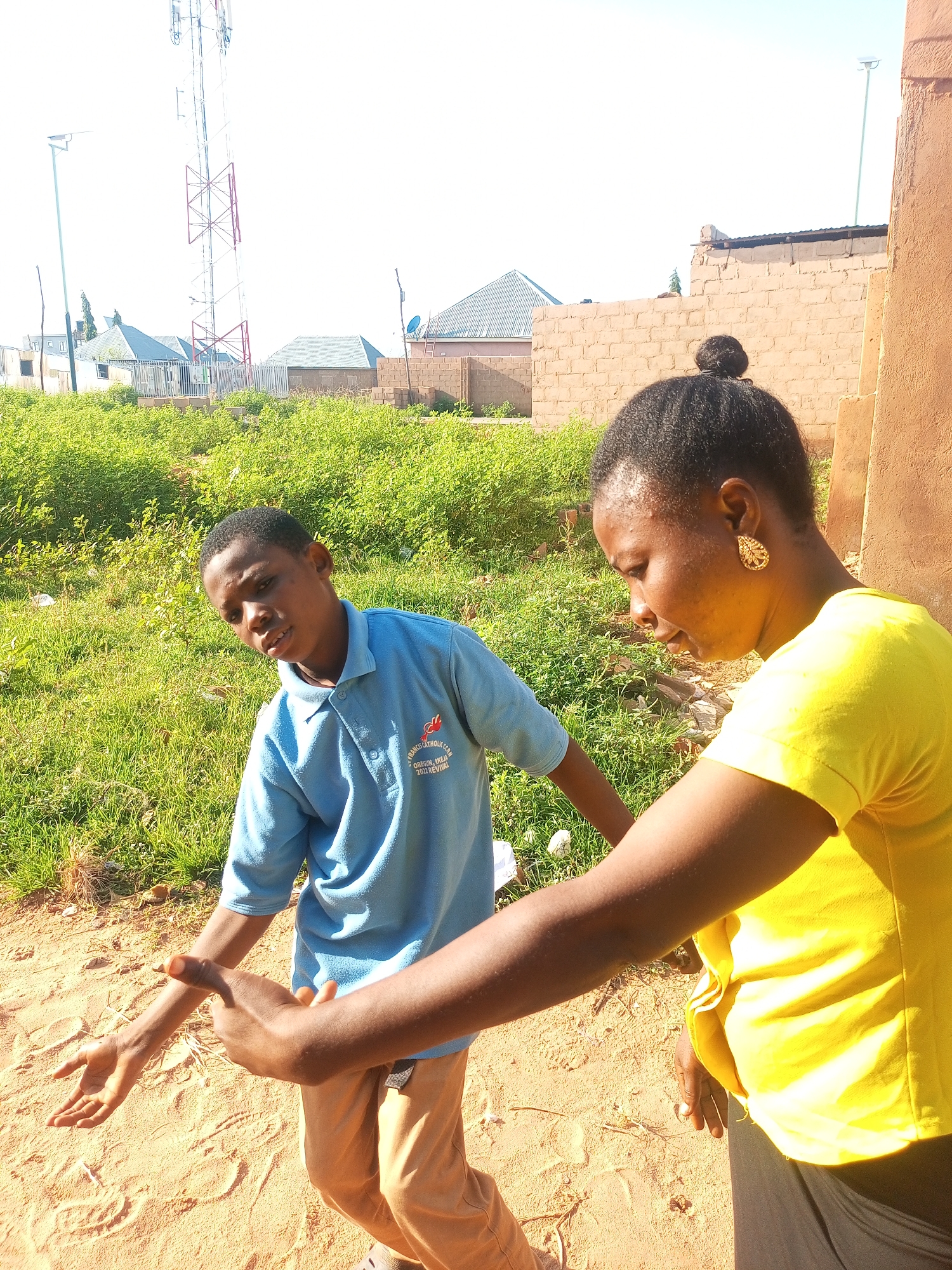 In a sunlit outdoor scene in the city of Tempure, Nigeria, a young boy in a light blue polo shirt and tan pants stands in sandy ground with patchy green grass and brush behind him. To his right, a woman in a bright yellow t-shirt and gray pants is looking down at his outstretched left arm, her right hand pointing towards his hand. In the background, a tall telecommunications tower with red and white bracing stands prominently, alongside several low-rise buildings with corrugated roofs, brick walls, and some construction materials. The sky is bright and clear.