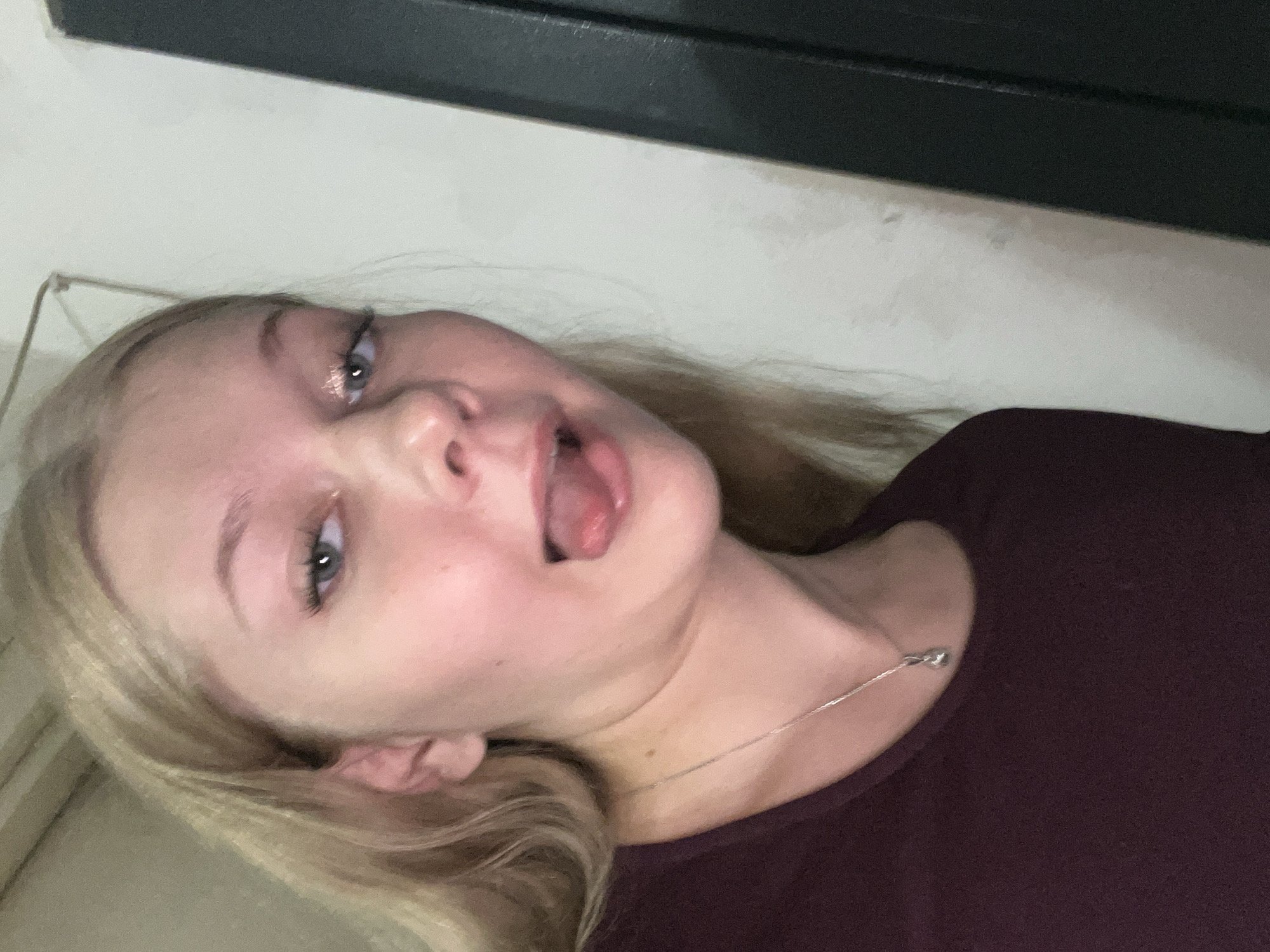 The image shows a close-up of a young woman with blonde hair, sticking her tongue out. She is wearing a dark maroon top and a silver necklace. The background is a plain wall, and a dark vertical object, possibly a door or furniture, is visible on the right side of the frame. The lighting suggests an indoor setting. There is no identifiable information to suggest a location in Perm, Russia.