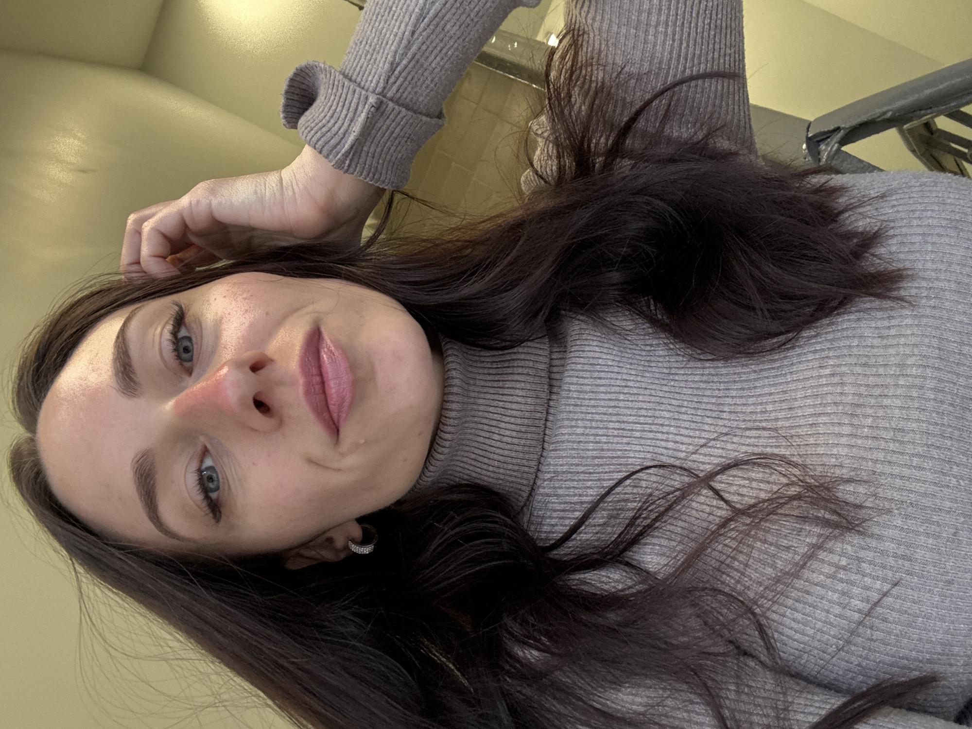 A selfie of a woman with long brown hair, light skin, and blue eyes. She is wearing a light grey turtleneck sweater and an earring. The background appears to be an indoor space, possibly a subway station, given the wall tiles and handrail visible. There is no definitive indication that this image was taken in St. Petersburg.