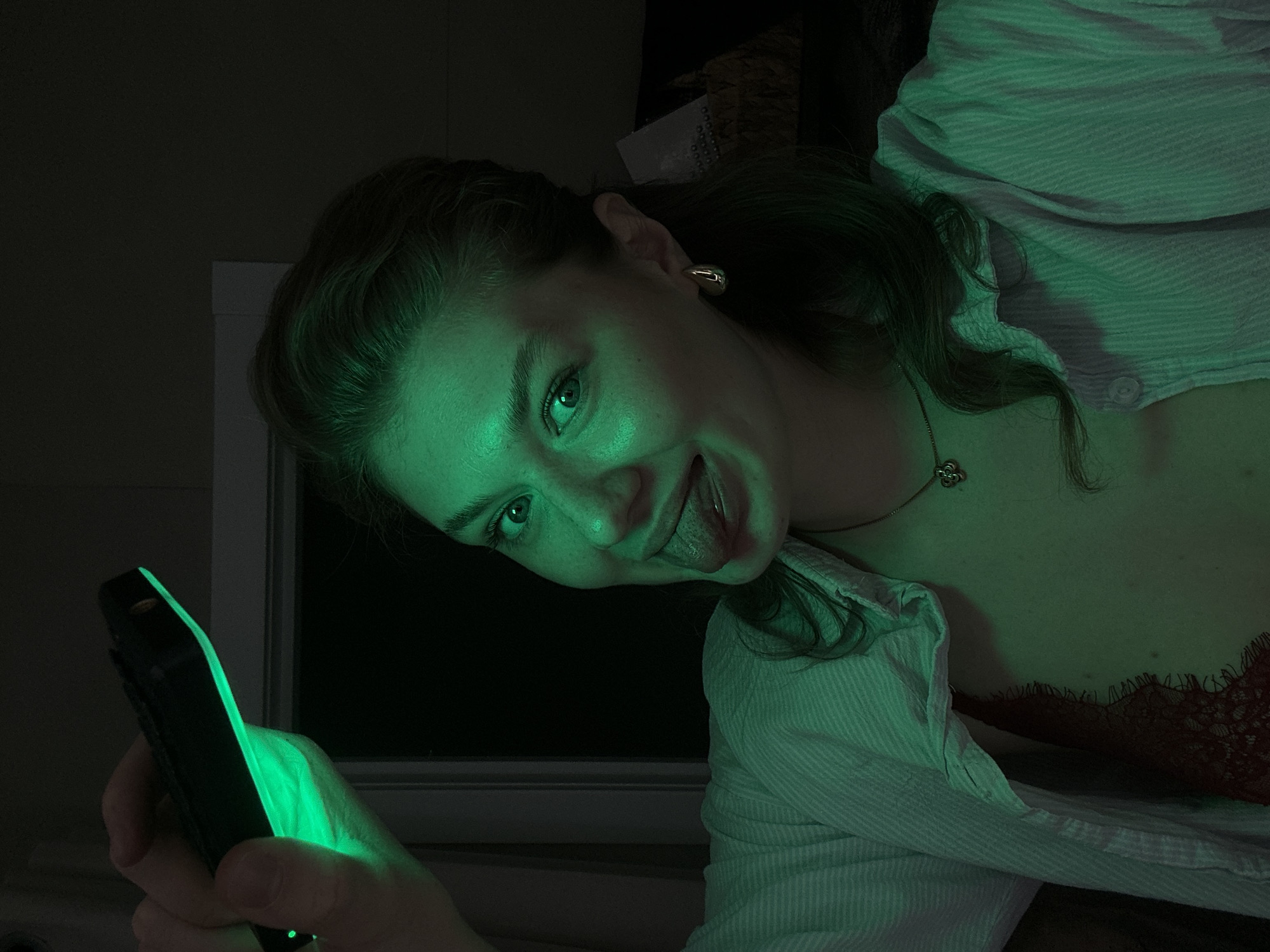 A woman is holding a smartphone that is emitting a green light. She is wearing a white striped shirt over a red lace top. She is sticking her tongue out. A window is behind her. The location is unknown.