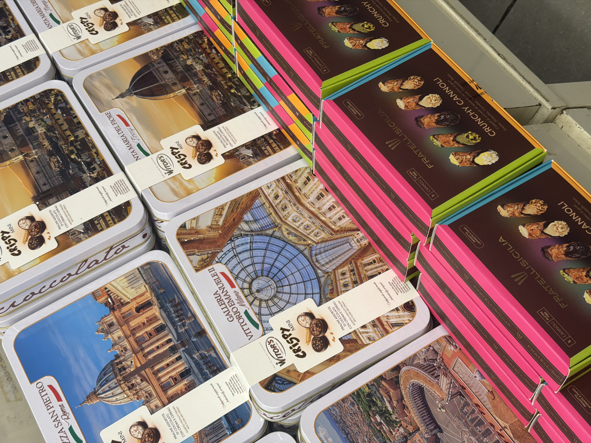 The image displays a retail display of assorted chocolate praline tins and cannoli boxes. In the foreground, several boxes of "FRATELLISICILIA CRUNCHY CANNOLI" are stacked, featuring vibrant pink and lime green packaging with images of cannoli. The background shows a variety of Witor's "CRISPY MINI" chocolate tins, each adorned with scenic images of famous Italian landmarks. One tin prominently features St. Peter's Basilica in Rome, another depicts the Duomo of Florence, and a third showcases the Galleria Vittorio Emanuele II in Milan. The overall impression is a product display in a shop or supermarket, likely in Italy given the Italian branding and landmarks. There is no discernible information in the image to suggest the location is specifically Ferrara, Italy; the depicted landmarks are from Rome, Florence, and Milan. The scene is well-lit and the products are neatly arranged. There are no people visible in the image.
