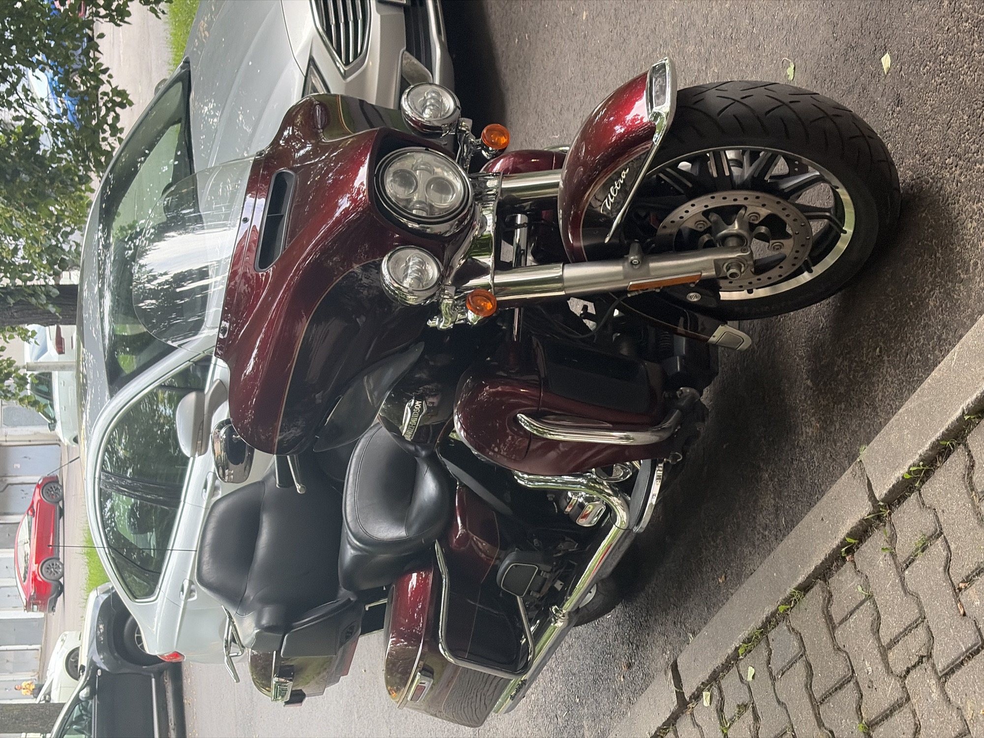 The image shows a red Harley-Davidson motorcycle parked on a street in Minsk, Belarus. The motorcycle is the primary subject and is viewed from the front right side. It has a large fairing, dual headlights, and chrome accents. The motorcycle is parked next to a silver car, and other cars are visible in the background. The street is paved with concrete blocks, and there are trees and foliage on the left side of the image. The sun is shining, casting shadows, suggesting it is daytime. There is no activity or event taking place, as the motorcycle is parked. Text visible on the motorcycle includes "Harley-Davidson" on the engine cover and "Ultra" on the fairing. The overall scene is a typical urban street view.
