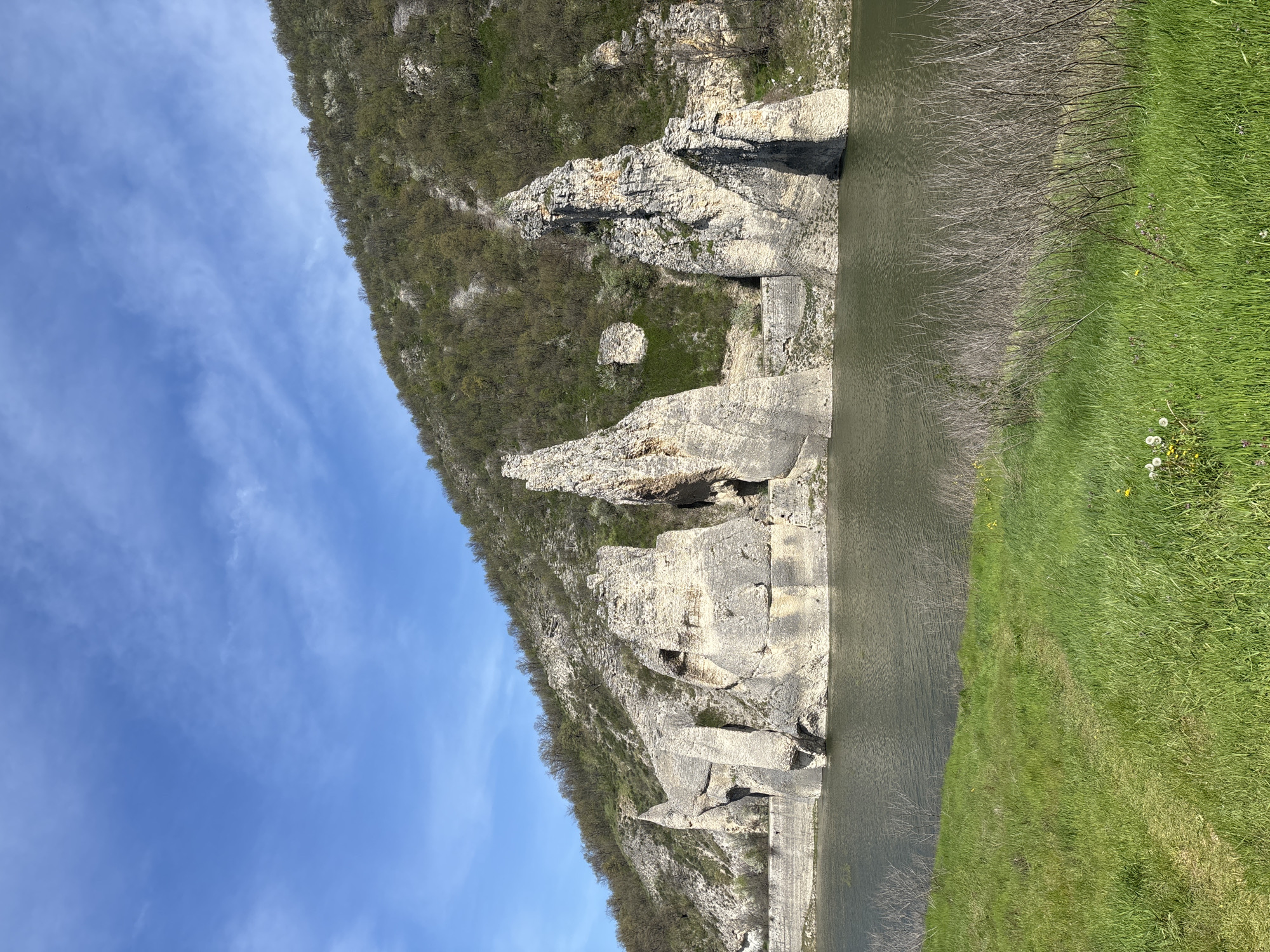 This media file captures a serene landscape featuring a body of water bordered by dramatic rock formations and lush greenery under a clear blue sky. The image appears to be taken during the daytime, likely in spring, given the fresh green foliage on the trees and grass.
The scene is dominated by a large expanse of water, which appears calm and reflects the sky and surrounding landscape. On the left side, towering rocky cliffs and formations rise sharply from the water's edge and extend upwards into a forested hillside. These light-colored rocks exhibit rugged textures and varied shapes, suggesting natural erosion over time. Some parts of the water's edge on this side also show what looks like a man-made concrete or stone structure. Further up the hillside, nestled among the trees, a small, block-like stone structure can be faintly seen, possibly ruins or a natural feature.
The hillsides beyond the rock formations are covered in dense vegetation, primarily deciduous trees and shrubs that are mostly bare but show early signs of green leaves, indicating the season. The right side of the image features a vibrant green grassy bank sloping down to the water. This grassy area is dotted with small yellow dandelions and other white wildflowers. At the immediate water's edge on the right, numerous dry, bare branches extend into the water, suggesting fluctuating water levels or overgrown shore vegetation.
The sky is a bright blue with scattered, wispy white clouds, indicating fair weather. No people or discernible activities are visible in the tranquil setting. The overall impression is one of natural beauty and peacefulness in the area of Asparuhovo, Bulgaria. There is no visible text.