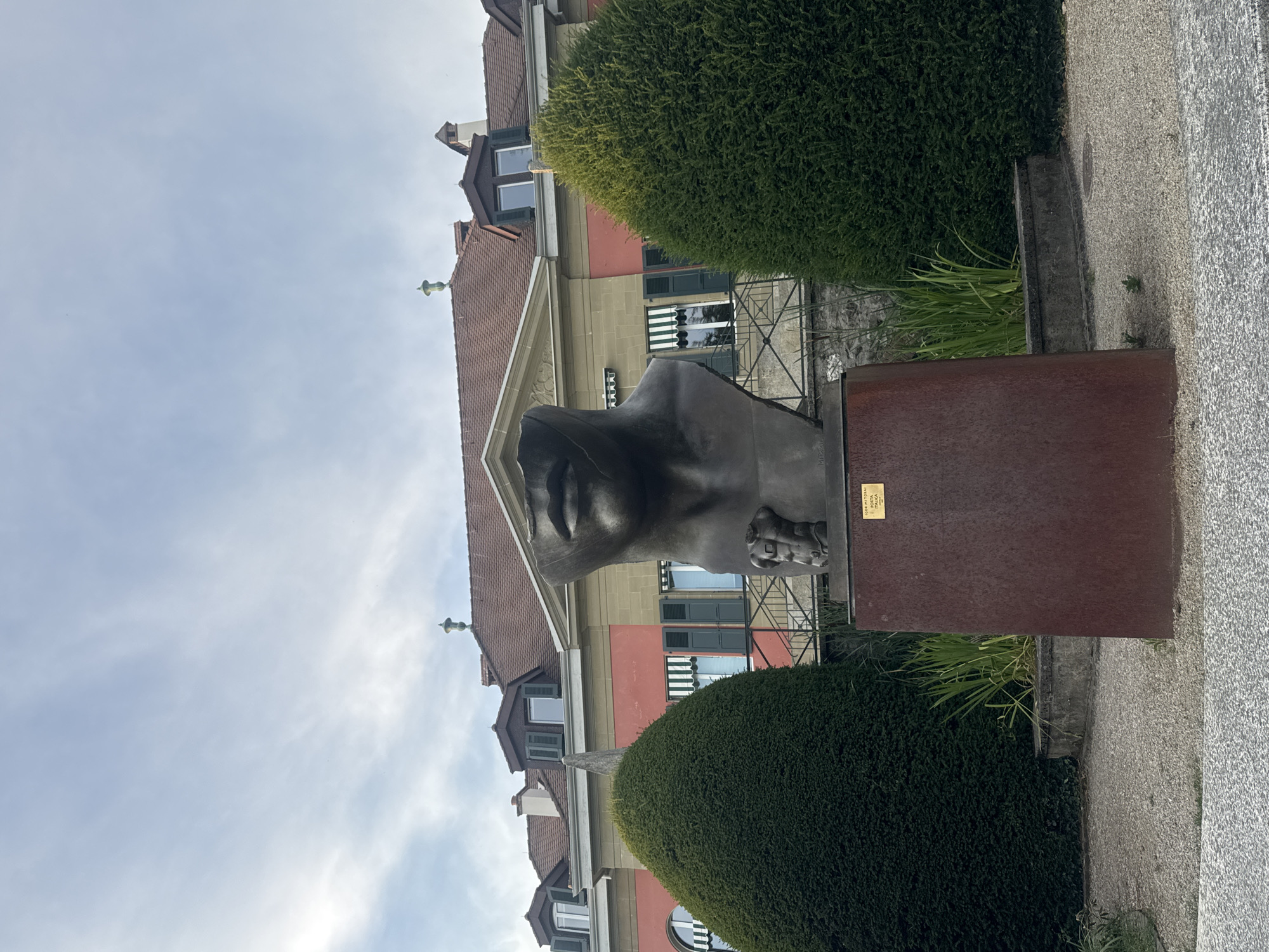 The photograph shows a large, abstract bronze sculpture of a head and torso with exposed musculature, positioned in front of a building. The sculpture is situated on a dark brown pedestal. To the left and right of the sculpture are two large, conical-shaped green bushes. Behind the sculpture and bushes is a building with a red roof, yellow and pink facades, and dark green shutters. The sky above is overcast with a light blue hue. The ground in front of the sculpture is covered with light-colored gravel. The sculpture appears to be located outdoors, in a public space such as a park or plaza. A small gold plaque is visible on the pedestal of the sculpture. The area is clean and well-maintained, suggesting an urban or semi-urban setting. This sculpture is known to be located in the Parc Mon-Repos in Lausanne, Switzerland, and is titled "Porta Italica" by Igor Mitoraj.