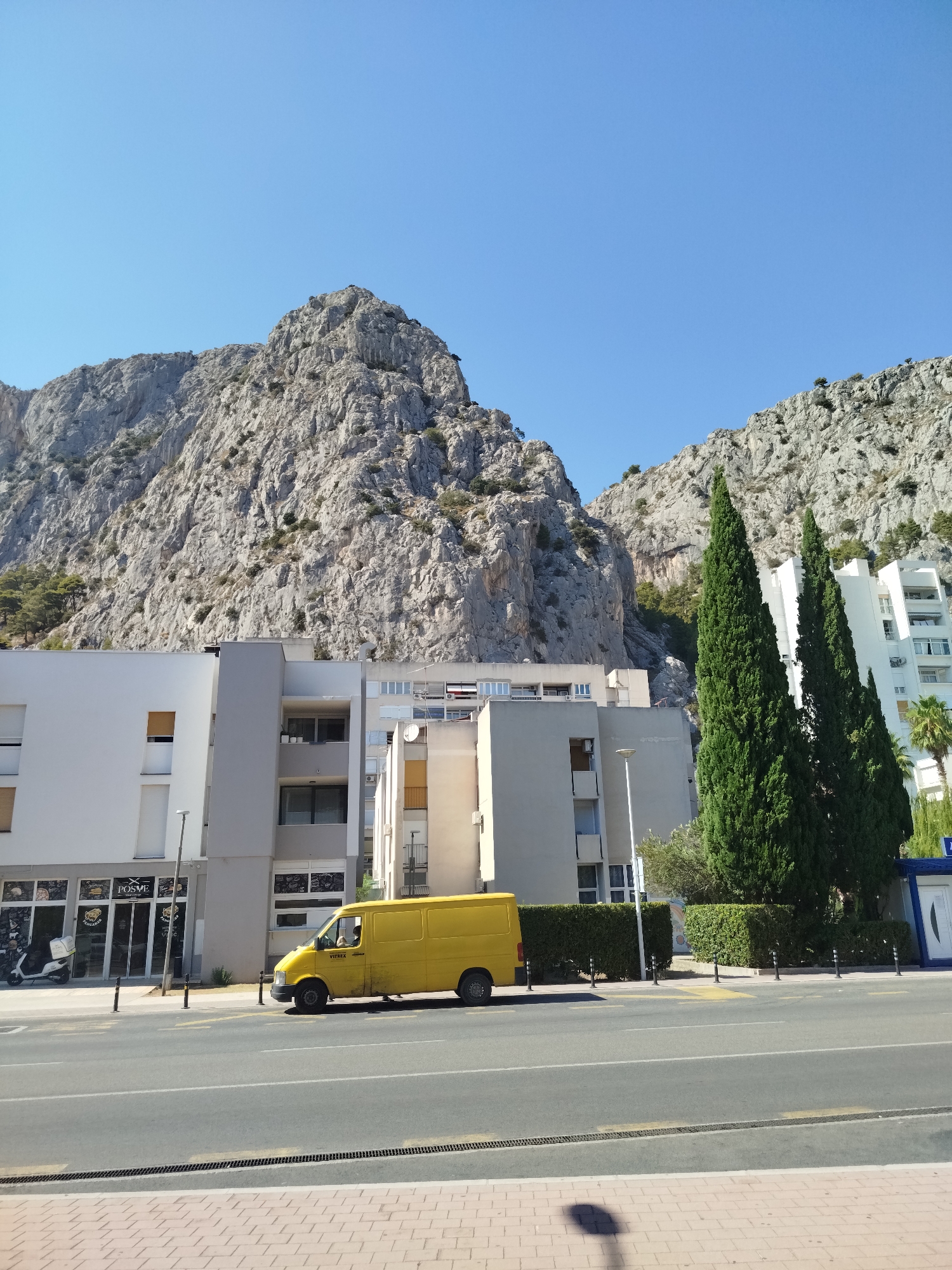 A bright yellow van is parked on a city street in front of a modern, light-colored building with several floors. The building is set against a backdrop of a large, rocky mountain range under a clear blue sky. Two tall, dark green cypress trees stand in front of the building, partially obscuring it. The street in the foreground is paved and has yellow lane markings. A sidewalk with brick paving is visible at the very bottom of the image. There are no visible people interacting, but a person can be seen inside the van through the driver's side window. The scene appears to be a sunny day in an urban environment adjacent to a natural mountain landscape.