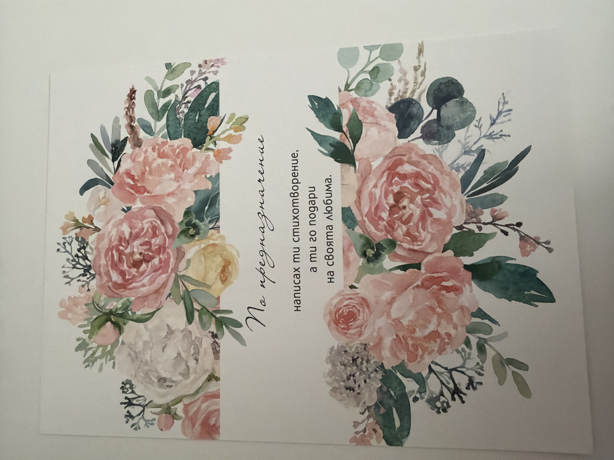 The image shows a close-up, eye-level, slightly angled, vertical shot of a white card with a floral design and text in Bulgarian. The floral design consists of watercolor illustrations of pink, white, and yellow roses, with green leaves and other foliage, arranged in two decorative borders at the top and bottom of the card. The text is handwritten in black ink. The top line reads "No предназначение" (No destination). Below this, in a smaller font, are the lines "написах ти стихотворение," (I wrote you a poem,) "а ти го подари" (and you gave it) "на своята любима." (to your beloved.) The background is a plain, off-white surface, likely a table or wall, which is out of focus. No specific location in Pazardzhik, Bulgaria is identifiable from this image.