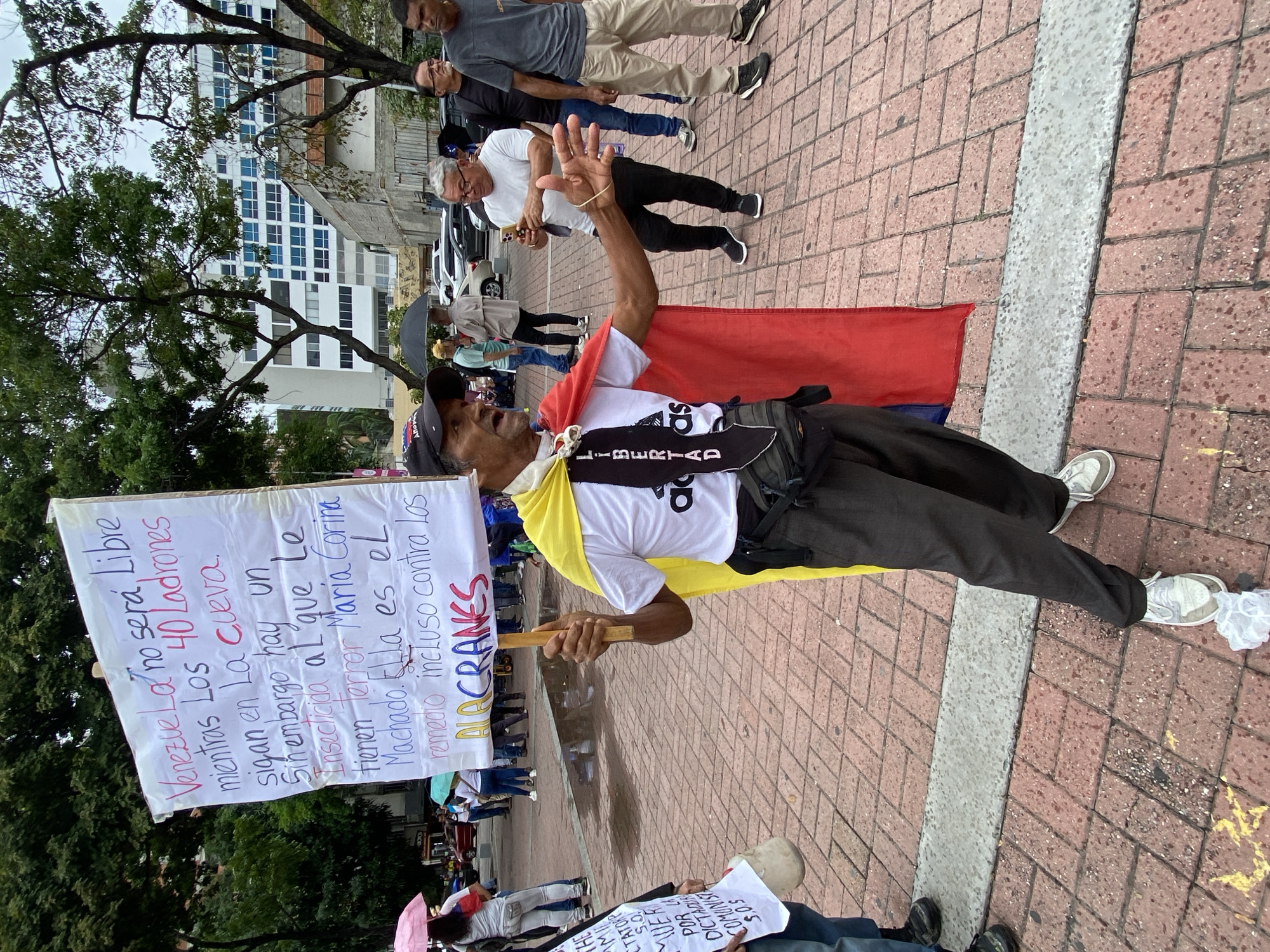 The image depicts an outdoor political protest taking place in Caracas, Venezuela. The scene is set on a paved walkway made of reddish-brown interlocking bricks, with a grey concrete strip running through it. The ground shows wet patches, indicating recent rain or an overcast day.
The central figure is a man, likely middle-aged or older, standing prominently with a large, handwritten protest sign. He wears a black baseball cap, a white t-shirt with partial text "LIBERTAD" visible, dark pants, and white sneakers. A red and yellow fabric is draped over his shoulders, reminiscent of the Venezuelan flag's colors. He holds the sign with both hands and has his right arm raised, palm open, as if making a statement or engaging with others.
The text on his sign, written in Spanish, reads: "Venezuela no será Libre mientras Los 40 Ladrones sigan en La cueva. Sin embargo hay un Insecticida al que le tienen Terror Maria Corina Machado. Ella es el remedio incluso contra los ALACRANES." This translates to: "Venezuela will not be free as long as the 40 thieves remain in the den. However, there is an insecticide that Maria Corina Machado terrifies. She is the remedy even against the SCORPIONS." This expresses strong support for opposition leader María Corina Machado and opposition to the current government.
In the background, other individuals are present, some holding umbrellas. Trees with green foliage frame the left side and background, while urban buildings, including a multi-story white building with blue-tinted windows, are visible further back. A smaller protest sign is partially visible in the lower right corner, stating "MUERTE POR LA DICTADURA COMUNISTA S.O.S LIBERTAD," meaning "DEATH BY THE COMMUNIST DICTATORSHIP S.O.S LIBERTY," reinforcing the anti-government sentiment of the gathering. The overall atmosphere is one of public dissent and a call for political change.