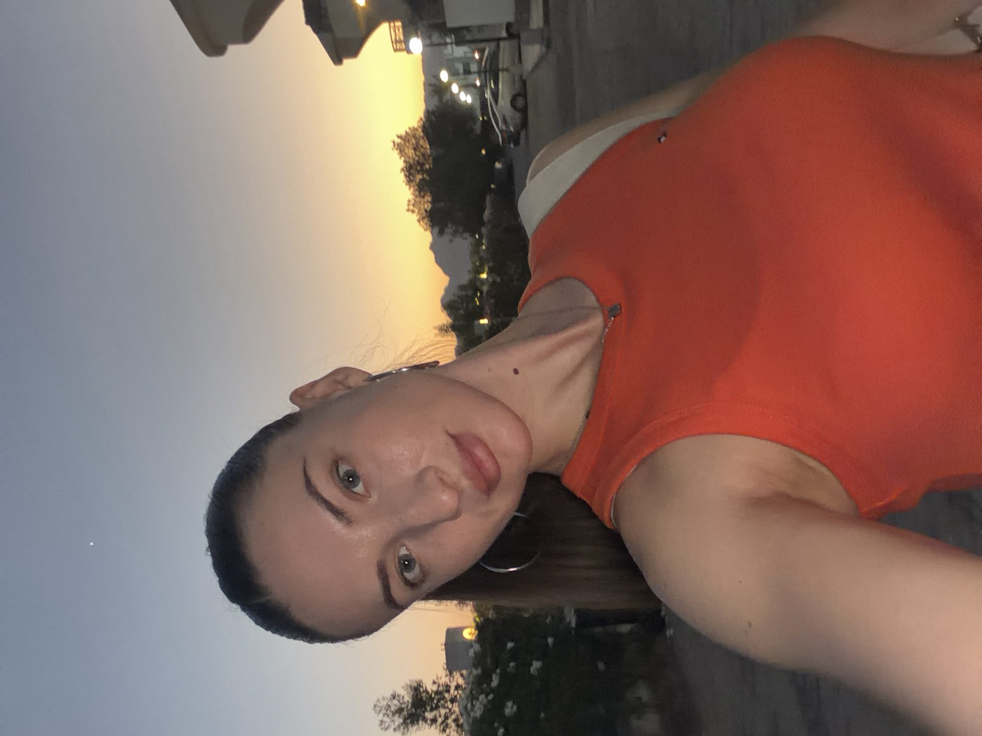 An outdoor, low-angle, selfie-style photograph captures a young woman posing against a backdrop of a twilight sky in Sharm El Sheikh, Egypt. The woman, with her hair pulled back in a ponytail and wearing a vibrant orange tank top and hoop earrings, looks directly into the camera. Her gaze is slightly to the left, and her head is tilted. In the background, a pale sky transitions from blue at the top to shades of orange and yellow near the horizon, with mountains visible in the distance. Streetlights and a few parked cars are also discernible, suggesting an urban or semi-urban setting at dusk. A white strap of a bag is visible over her shoulder. The overall ambiance suggests a casual moment captured during the evening in a warm climate.