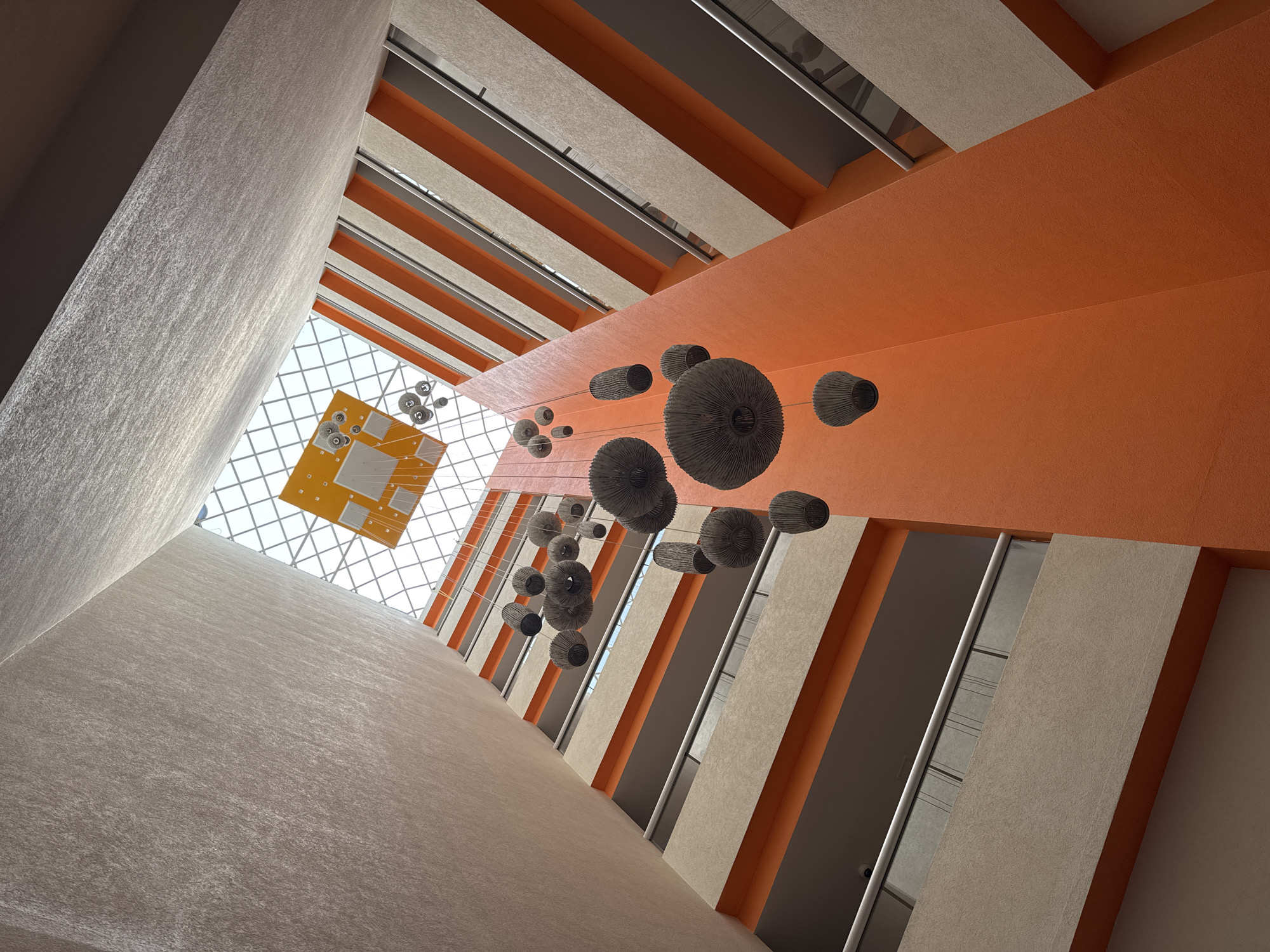 The image shows an upward view of an interior building space in Querétaro, Mexico. The building has a multi-story atrium with alternating bands of beige and orange walls. A series of decorative, spherical light fixtures are suspended within the atrium space. A yellow, rectangular structure is visible at the top of the atrium through a grid-patterned skylight, along with other smaller round light fixtures. The space appears modern and architectural in design. The walls have a textured surface. The view is angled significantly upwards.