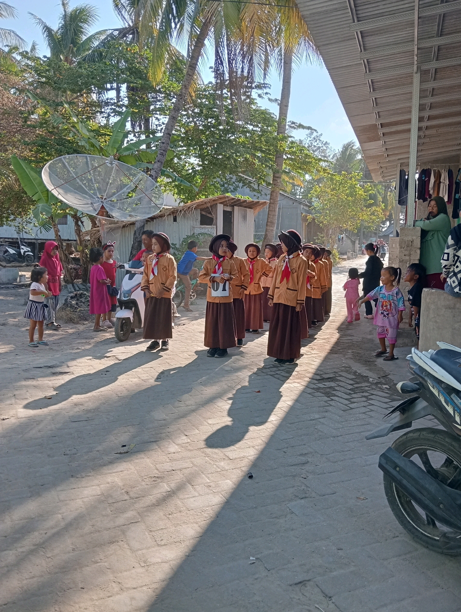 In Pagerungan Kecil, Indonesia, under a clear sky, a group of children in uniform stand in a line on a paved street. The children appear to be participating in an event, with some wearing matching brown uniforms and hats, suggesting a school or scouting activity. One child in the foreground wears a brown uniform with a red and yellow sash, and a number "01" is visible on their uniform. Another child to their right wears a similar uniform without a visible number. Behind them, other children in similar attire are also lined up, their faces turned forward.
To the left of the main group, a younger child in a pink shirt and striped skirt stands facing the line. Further back on the left, a child in a red headscarf and a pink dress stands with another child in a pink dress. A white scooter is parked to the left of the children in uniform.
The environment is a street with houses and buildings lining one side, some with corrugated metal roofs. Palm trees and other lush greenery are visible in the background, indicating a tropical setting. A large satellite dish is mounted on a pole to the left of the scene. On the right side of the street, a woman in a green headscarf stands near a building that appears to be a shop or market, with clothes displayed on a rack. Another woman in a patterned top stands to her right. A motorcycle is parked on the right side of the street in the foreground, its rear wheel and exhaust pipe visible. The sun casts long shadows of the people and objects onto the street. The overall scene depicts a community gathering or event involving children.