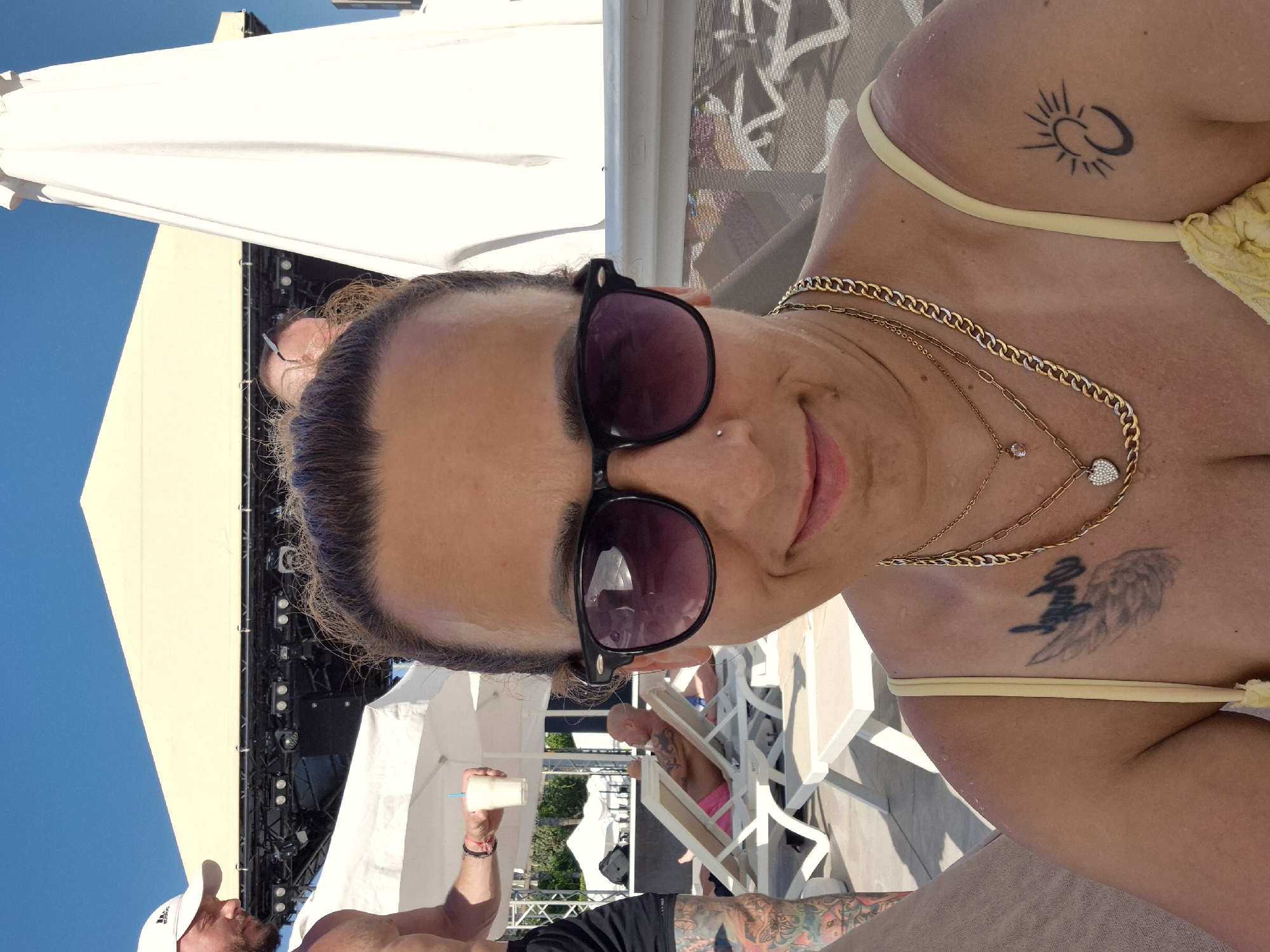 This is a close-up, low-angle, vertical photograph of a woman in a yellow bikini, holding a drink. She is wearing sunglasses and has multiple necklaces. There are two visible tattoos on her upper body: a sun-like design on her shoulder and an angel wing design on her chest. She appears to be smiling and looking at the camera.
In the background, a man wearing a white baseball cap is partially visible, holding a drink. Further in the background, other people are seen relaxing on lounge chairs under a bright blue sky, suggesting an outdoor, daytime setting. A large white structure, possibly a tent or canopy, is also visible. The overall impression is a casual, sunny day, likely at a resort or pool area. There are no discernible text elements in the image.