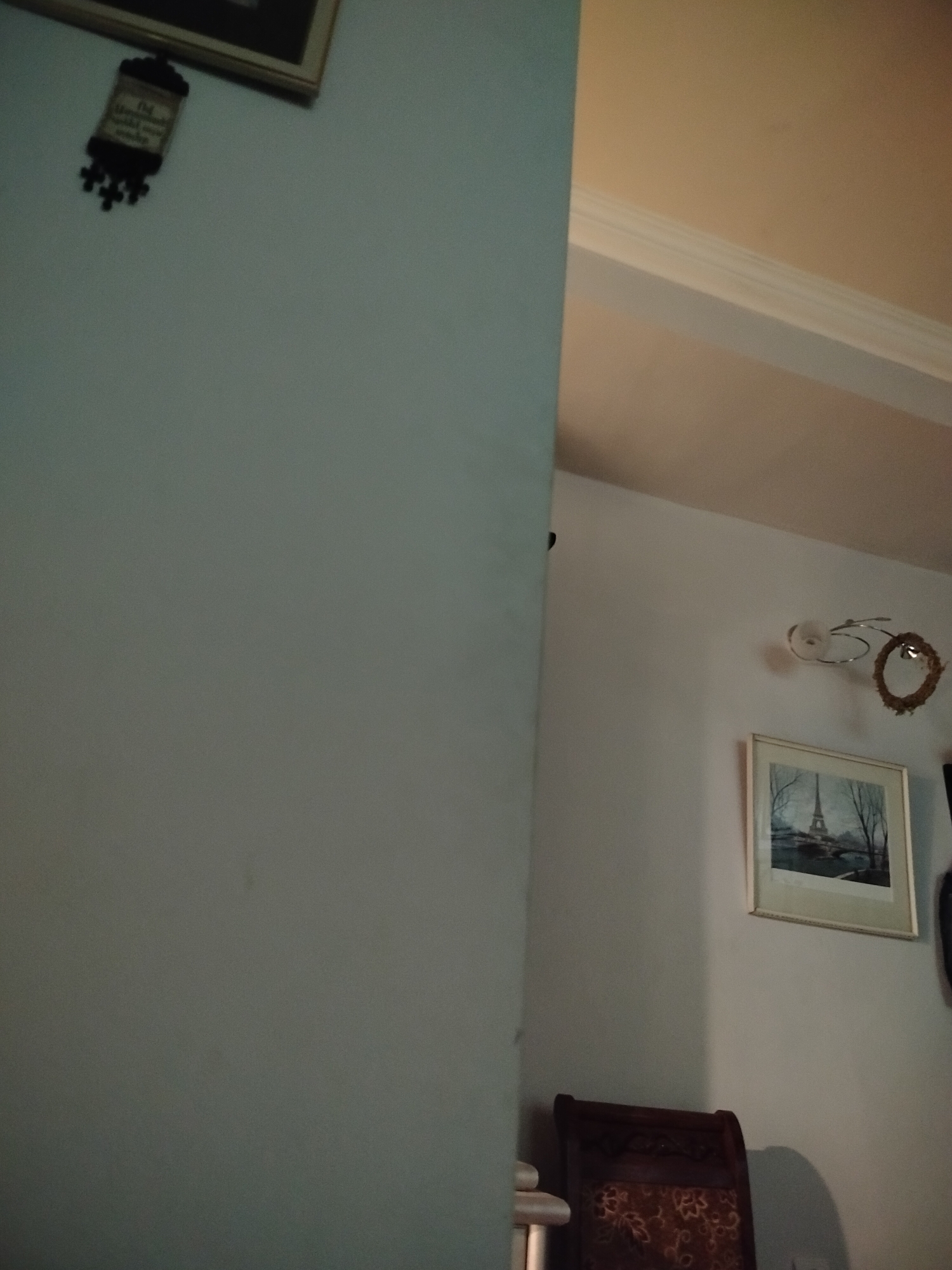 The image shows an interior corner of a room, with a light blue wall on the left and a white wall on the right. On the left wall, there is a framed artwork and a smaller decorative object. On the right wall, there's a framed picture of the Eiffel Tower, a light fixture, a decorative wreath, and a chair. The location in Yerevan, Armenia, is not discernible from the image.