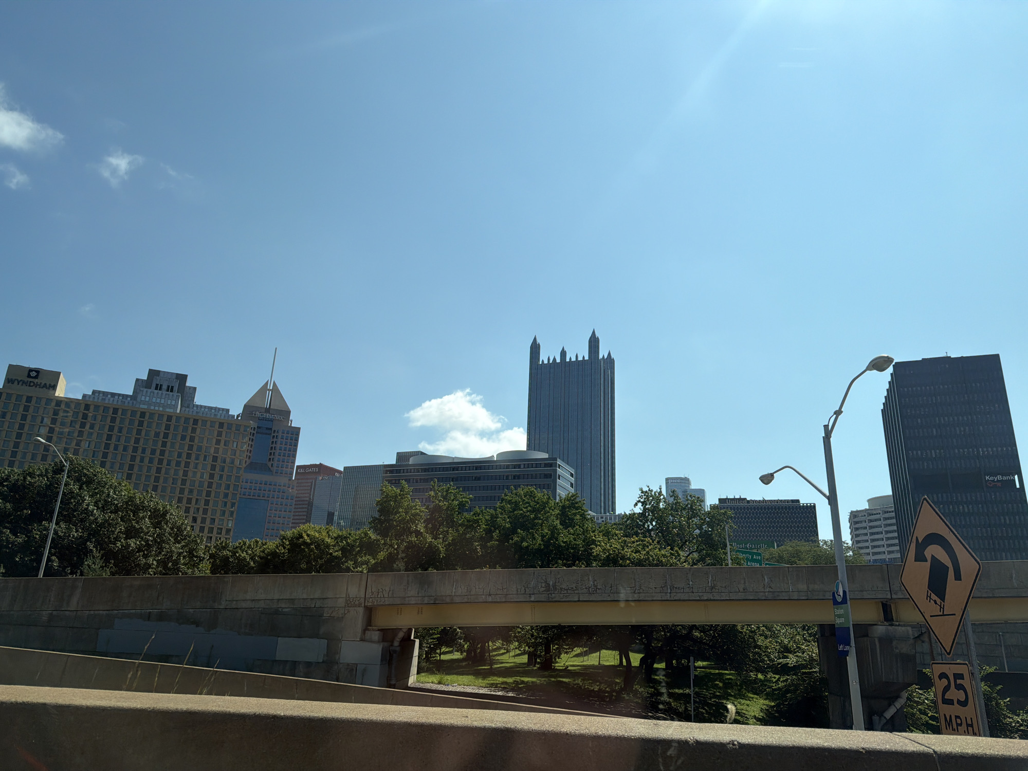 The image captures a sunny day in Pittsburgh, Pennsylvania, with a clear blue sky and a few scattered clouds. The foreground is dominated by a concrete barrier, suggesting the perspective is from a vehicle on an elevated road or highway. A yellow beam supports an overpass structure, with greenery and trees visible beneath it.
In the background, the Pittsburgh skyline is visible, featuring a collection of modern and older skyscrapers. Notable buildings include the distinctive U.S. Steel Tower with its unique top, and the dark, angular KeyBank building. Other buildings of varying heights and architectural styles populate the urban landscape. Several road signs are present, including a diamond-shaped directional sign and a speed limit sign indicating "25 M.P.H." A lamppost with a curved arm stands near the KeyBank building. The overall scene depicts a typical urban view of Pittsburgh's downtown area from a highway perspective.