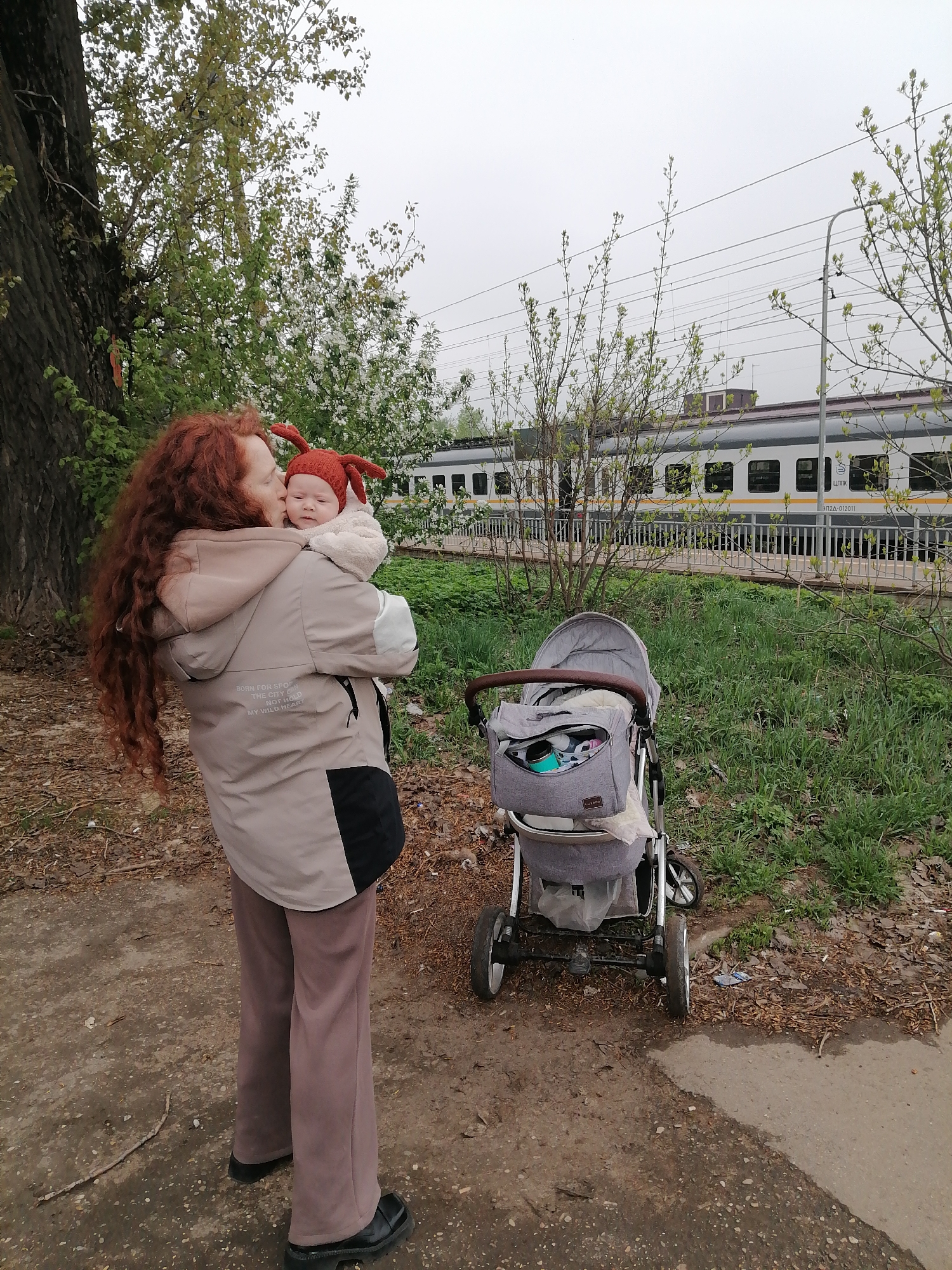 A woman with long red hair holds a baby in a red hat and kisses the baby's forehead. The woman is wearing a beige jacket, and the baby is wearing a white outfit. Next to them is a gray baby stroller. Behind them is a train and some vegetation. The photo was taken in Юдино, Russia.