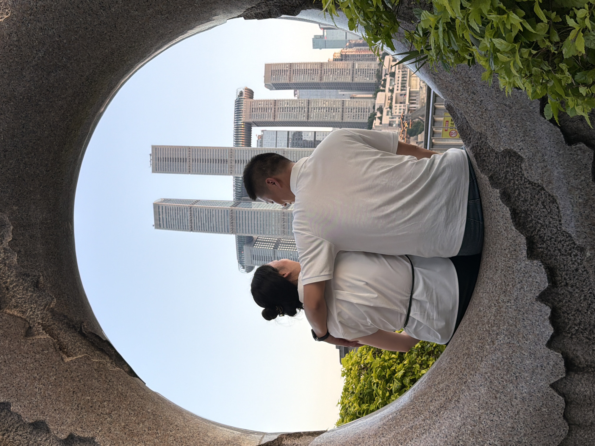 A couple sits on a stone bench, framed by a circular stone opening. The man has his arm around the woman's shoulders as they both gaze at a cityscape. The woman has her hair in a bun and is wearing a white top and dark pants. The man is wearing a white polo shirt and blue jeans. In the background, modern skyscrapers dominate the skyline, with the distinctive Raffles City Chongqing complex, featuring twin towers connected by a horizontal skybridge, prominently visible. The scene is set in the city of Nan'an District, China, with lush green foliage in the foreground, partially obscuring the view. The sky is clear and blue, indicating it is daytime.