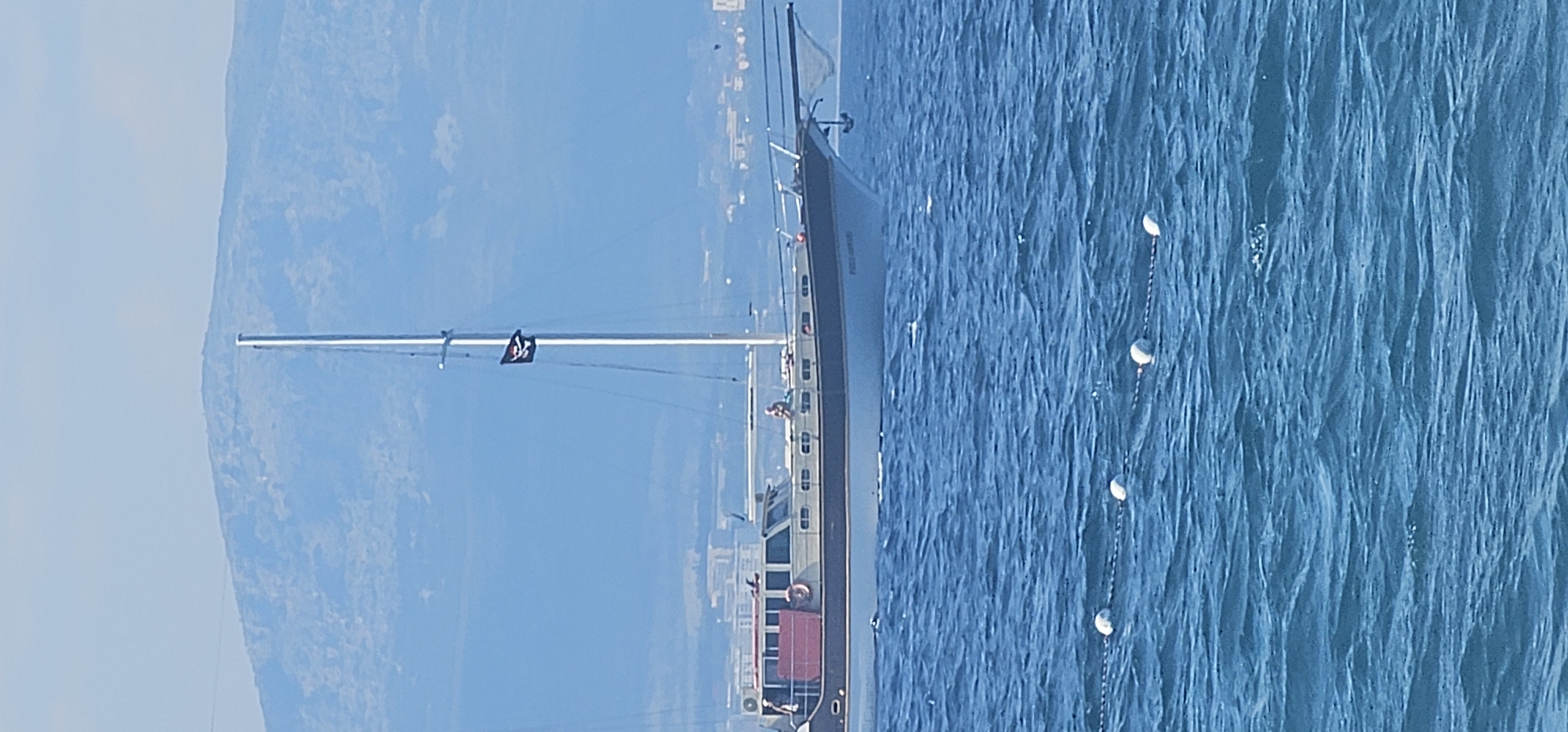 In the city of Supetar, Croatia, on a bright day, a large white sailboat with a brown trim sails on the blue sea. The sailboat has a tall mast with a black pirate flag with a skull and crossbones. There is a person visible on the deck of the sailboat. In the background, a hazy blue mountain range rises above the distant city skyline. The foreground shows the rippling surface of the ocean, with a chain link floating device marking the water.