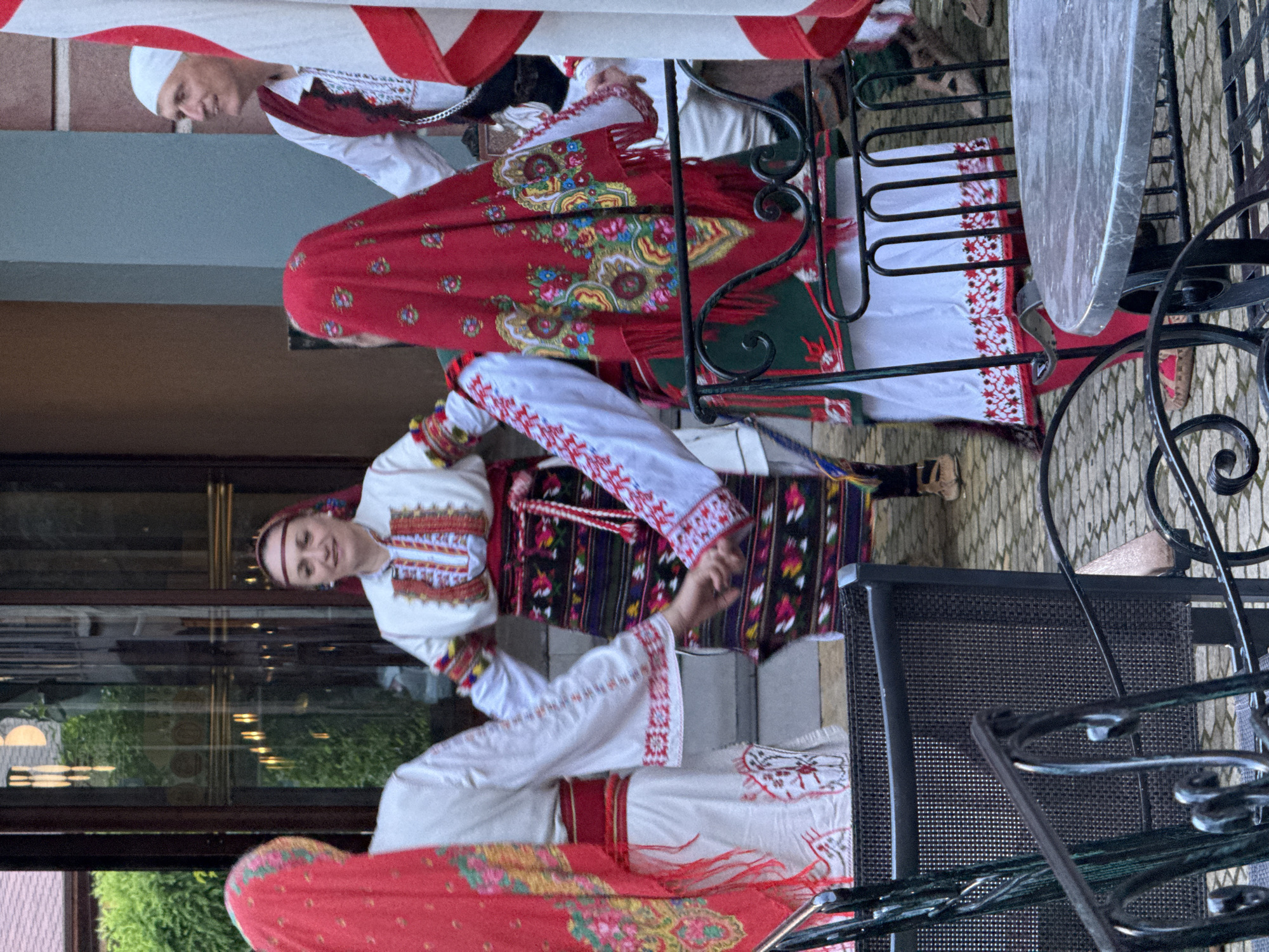 In Kyustendil, Bulgaria, a group of four people are dancing in a circle, holding hands. The two individuals in the foreground are wearing traditional Bulgarian attire. The woman on the left has a red headscarf with floral embroidery and a white dress with red trim. The woman on the right is wearing a red headscarf with an intricate floral pattern, a white blouse with colorful embroidered sleeves, and a dark skirt with horizontal stripes. Her hands are clasped with the person next to her. To the right, a man is visible wearing a white shirt, a red vest, and a white head covering. He is smiling and looking to his right. The background features a building with glass doors and green foliage. In the foreground, there are chairs and a table, suggesting an outdoor seating area. The ground is paved with cobblestones.
