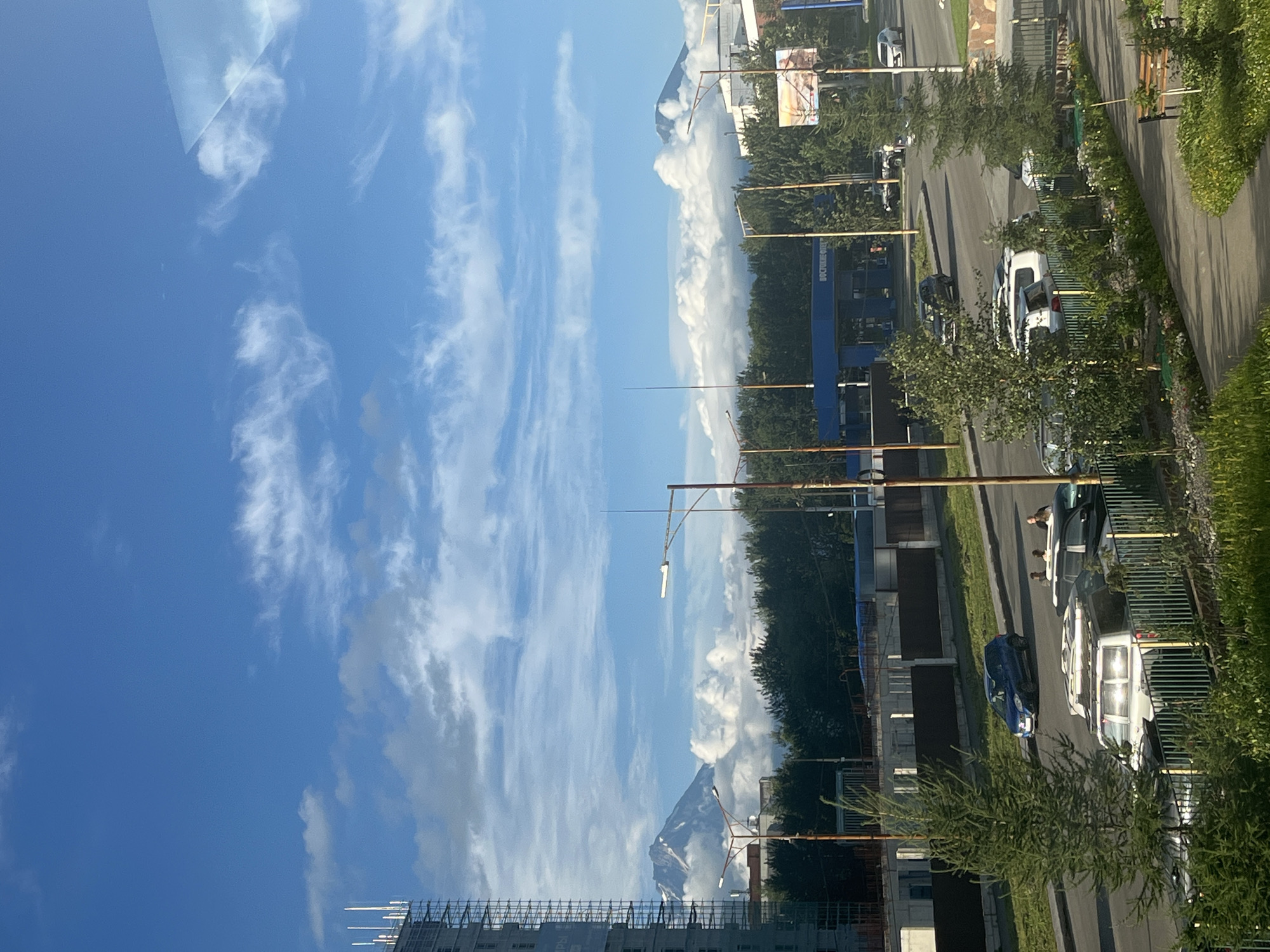 The image captures a street scene in Petropavlovsk-Kamchatsky, Russia, dominated by a bright blue sky with scattered white clouds. In the background, two majestic, snow-capped volcanoes are visible, partially obscured by fluffy white clouds that rise from behind a dense line of green trees. To the left, a tall building under construction, clad in scaffolding, stands prominently.
In the mid-ground, a road runs horizontally, with several cars parked and in motion. A blue SUV is driving on the road, while various other vehicles, including what appear to be white SUVs and a black sedan, are parked along the roadside. Some people are visible near the parked vehicles. A blue building with a sign that reads "ВОСТОЧНАЯ" (Vostochnaya) is situated near the road, appearing to be a gas station or convenience store. Streetlights with multiple lamps are strategically placed along the road.
The foreground features a paved walkway, a neatly trimmed green lawn with flowerbeds, and a wooden bench. A tall evergreen tree is positioned to the left of the walkway, and a smaller pine tree is closer to the road. A green metal fence separates the walkway and lawn area from the street. The overall impression is a juxtaposition of urban development and the impressive natural landscape of the region.