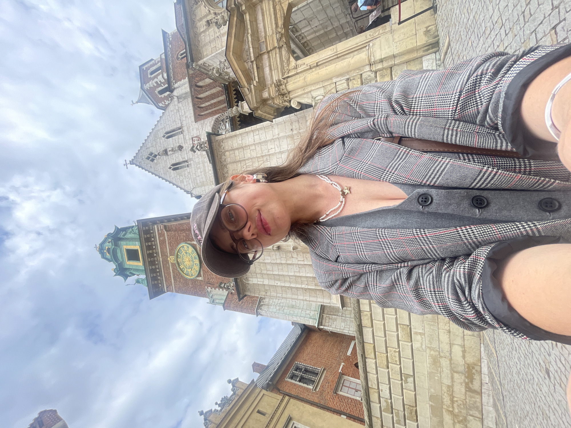 The image shows a young woman taking a selfie in front of Wawel Castle in Krakow, Poland. She is wearing a grey plaid blazer over a grey vest and a cap. She has glasses and is wearing a pearl necklace with a star pendant. In the background, the clock tower of Wawel Cathedral is visible. The sky is cloudy. The woman is holding a phone in her hand to take the selfie.
