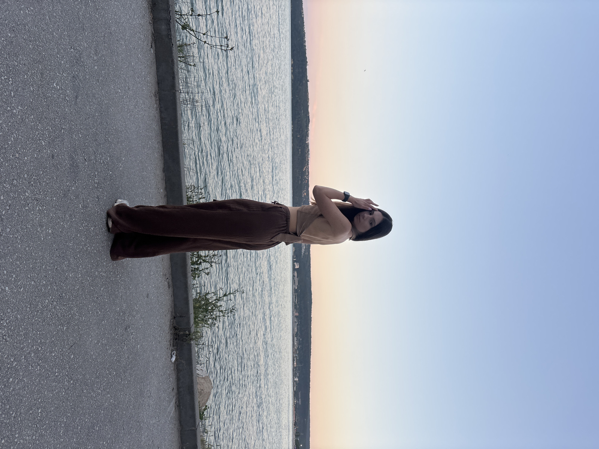 A young woman with long, dark hair stands on an asphalt pathway next to a body of water, likely the Black Sea, with a coastal city visible in the distance. The woman is wearing a beige halter top and wide-leg brown pants. She is looking over her left shoulder towards the camera, with her right hand touching her hair. The sky is a gradient of light blue and pale pink, indicating either sunrise or sunset. The water is calm with gentle ripples, and a line of concrete blocks separates the pathway from a narrow strip of vegetation and a rock. The city in the background appears to be Varna, Bulgaria, with buildings and structures visible along the coastline.