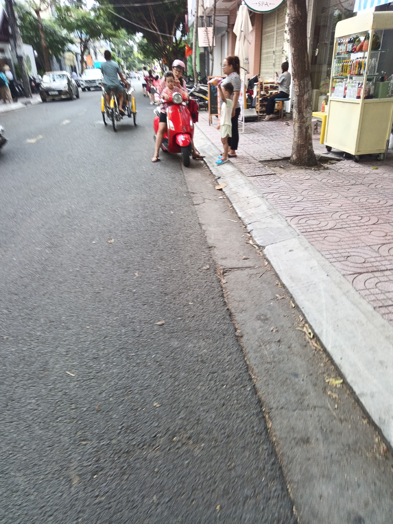 In Nha Trang, Vietnam, a street scene unfolds with a prominent red scooter in the foreground, carrying a woman and a child. The woman, wearing a pink helmet, is positioned behind the child, who is seated on the scooter. To the left of the scooter, a woman in a striped shirt stands with a young child wearing a white t-shirt and shorts. Behind them, a vendor's cart filled with colorful drinks and fruit is visible. Further down the street, traffic, including cars and other scooters, is present. The environment includes trees lining the street and a sidewalk with patterned tiles. The overall image captures a slice of daily life on a busy street in this Vietnamese city.