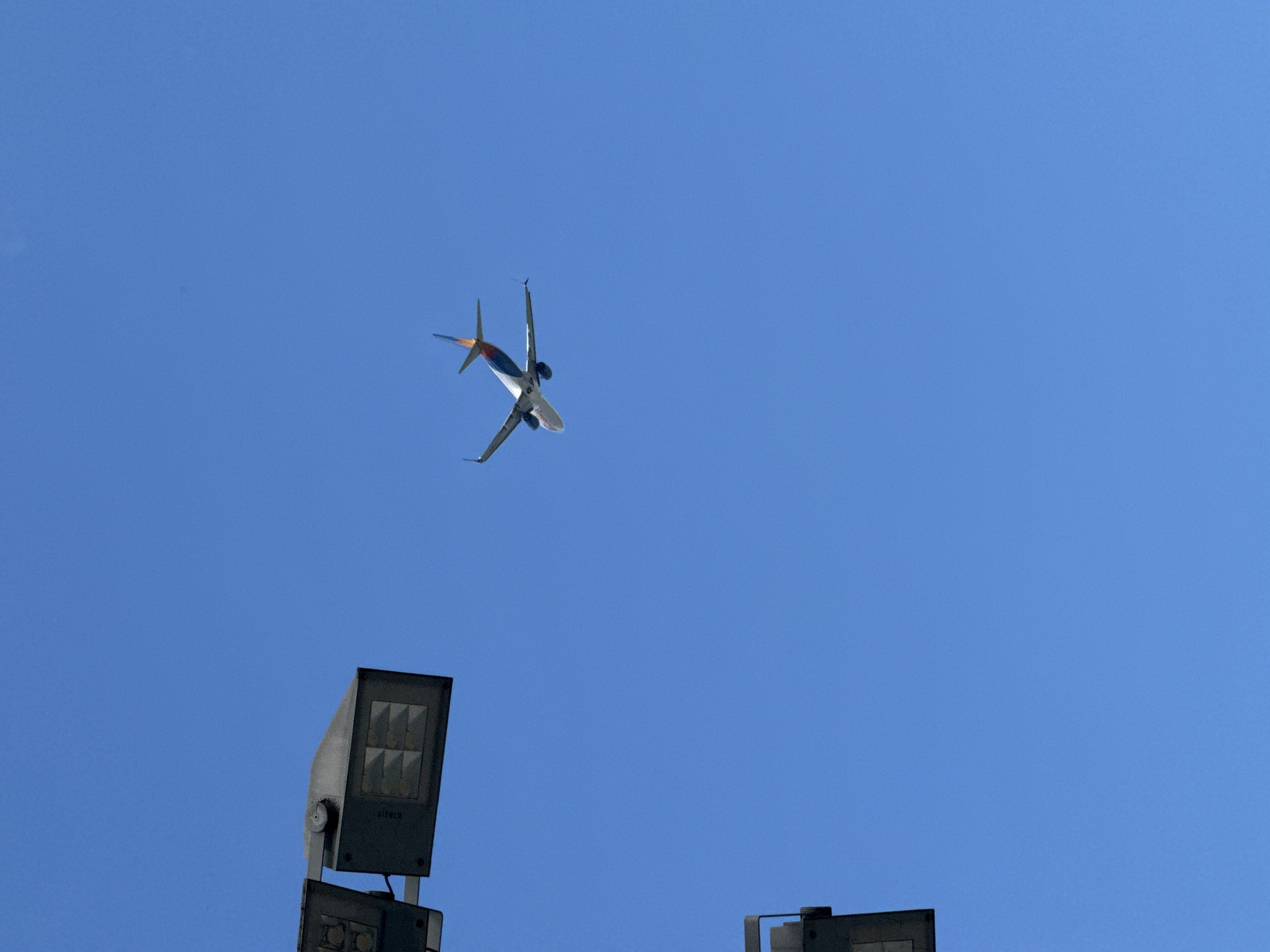 A commercial airplane with a white fuselage and a colorful tail is captured in mid-flight against a clear blue sky. The plane's wings are angled upwards, suggesting it is either ascending or banking. In the foreground, partially visible, are parts of two large outdoor light fixtures, suggesting the image was taken from a location with elevated lighting, possibly an airport or a stadium. The overall scene is devoid of any specific landmarks that would definitively place it in Innsbruck, Austria.