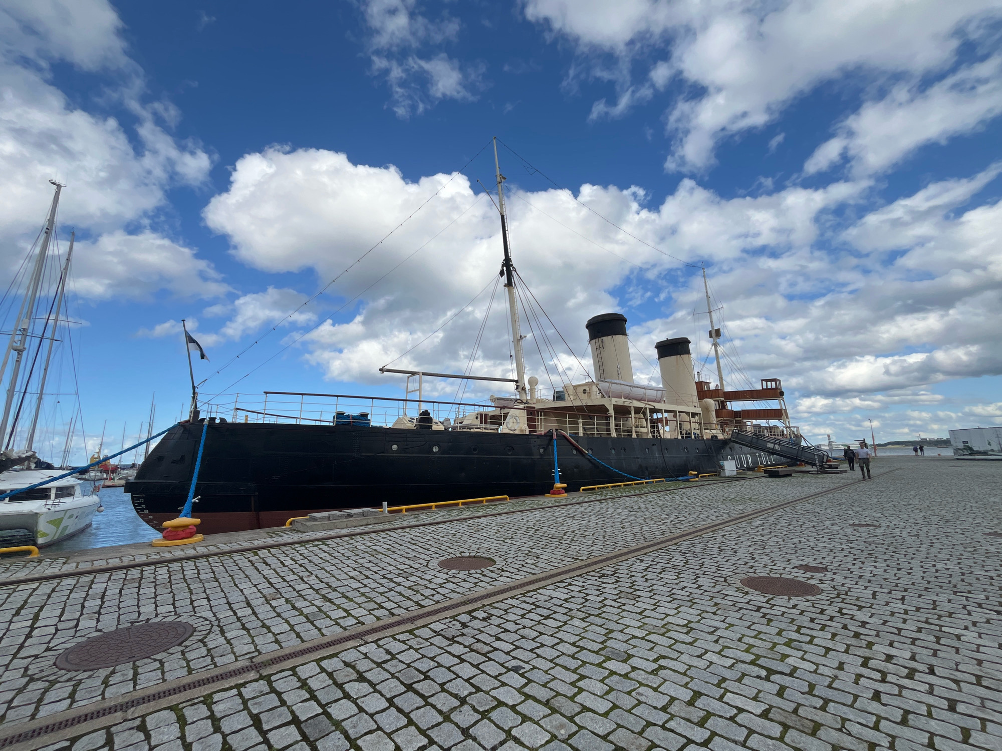 A large, black steamship is docked at a cobblestone pier under a blue sky with scattered white clouds. The ship, with its tall smokestacks and wooden superstructure, appears to be a historical vessel. A gangway labeled "JUUR TOLL" is extended from the ship to the pier. Several sailboats are visible in the background, moored in the harbor. Two individuals are walking on the pier, one in the foreground and another further away near the ship. The cobblestone pavement is well-maintained, with some metal grates and manhole covers integrated into its surface. The scene suggests a port or maritime museum in Tallinn, Estonia, given the ship's appearance and the label on the gangway.