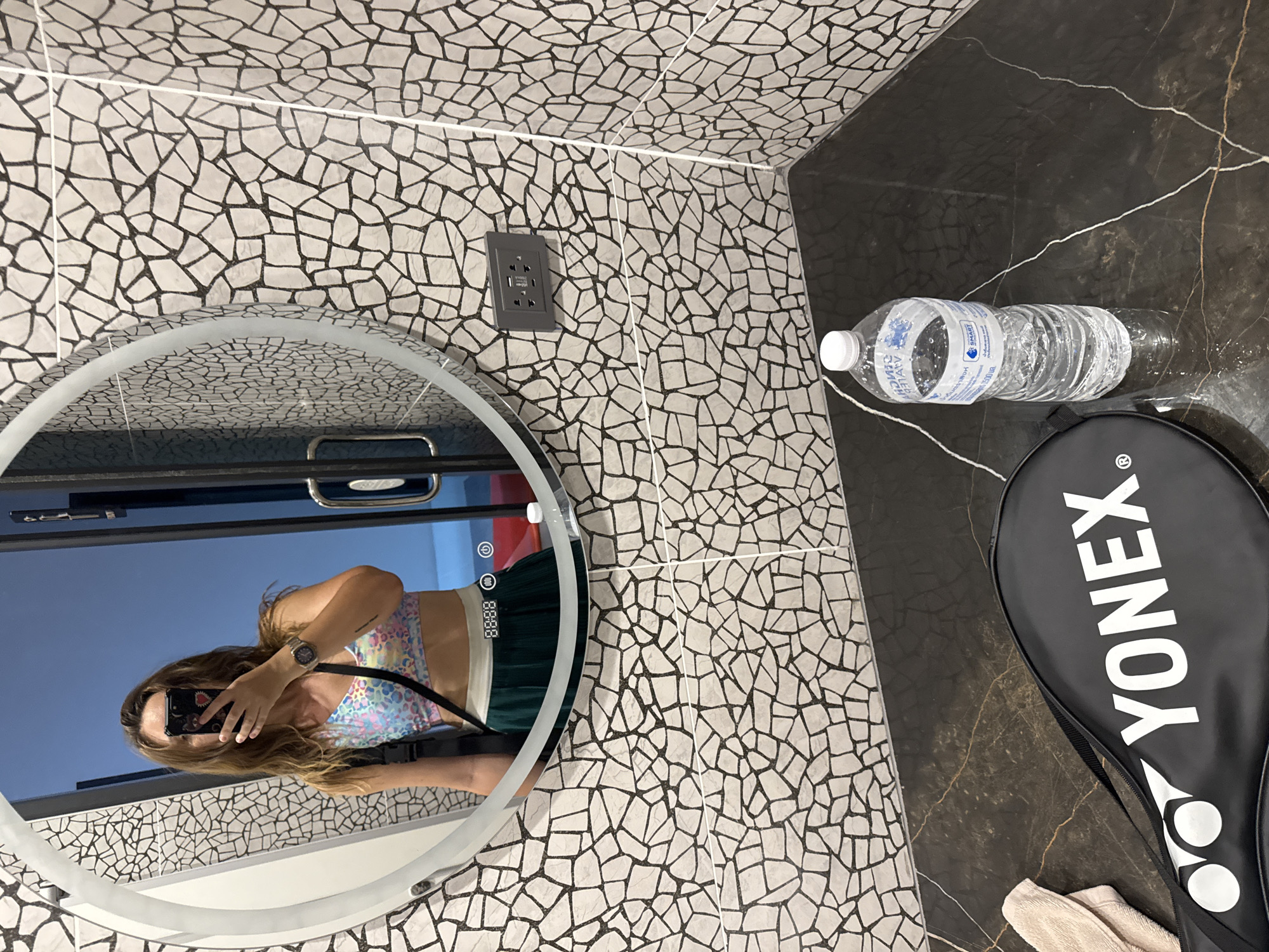 A woman is taking a selfie in a bathroom mirror. She is wearing a colorful, leopard-print athletic top and a green skirt. She has long, light brown hair and is wearing a watch on her left wrist. The bathroom has a unique tile pattern on the walls, featuring irregular shapes in black and white. A large, round mirror is mounted on the wall. Next to the mirror, a power outlet is visible. To the right of the mirror, a bottle of water and a black Yonex tennis racket bag are placed on a dark, marble-like countertop. The overall impression is that of a modern, stylish bathroom. The lighting suggests it could be daytime or artificial lighting in an indoor setting.