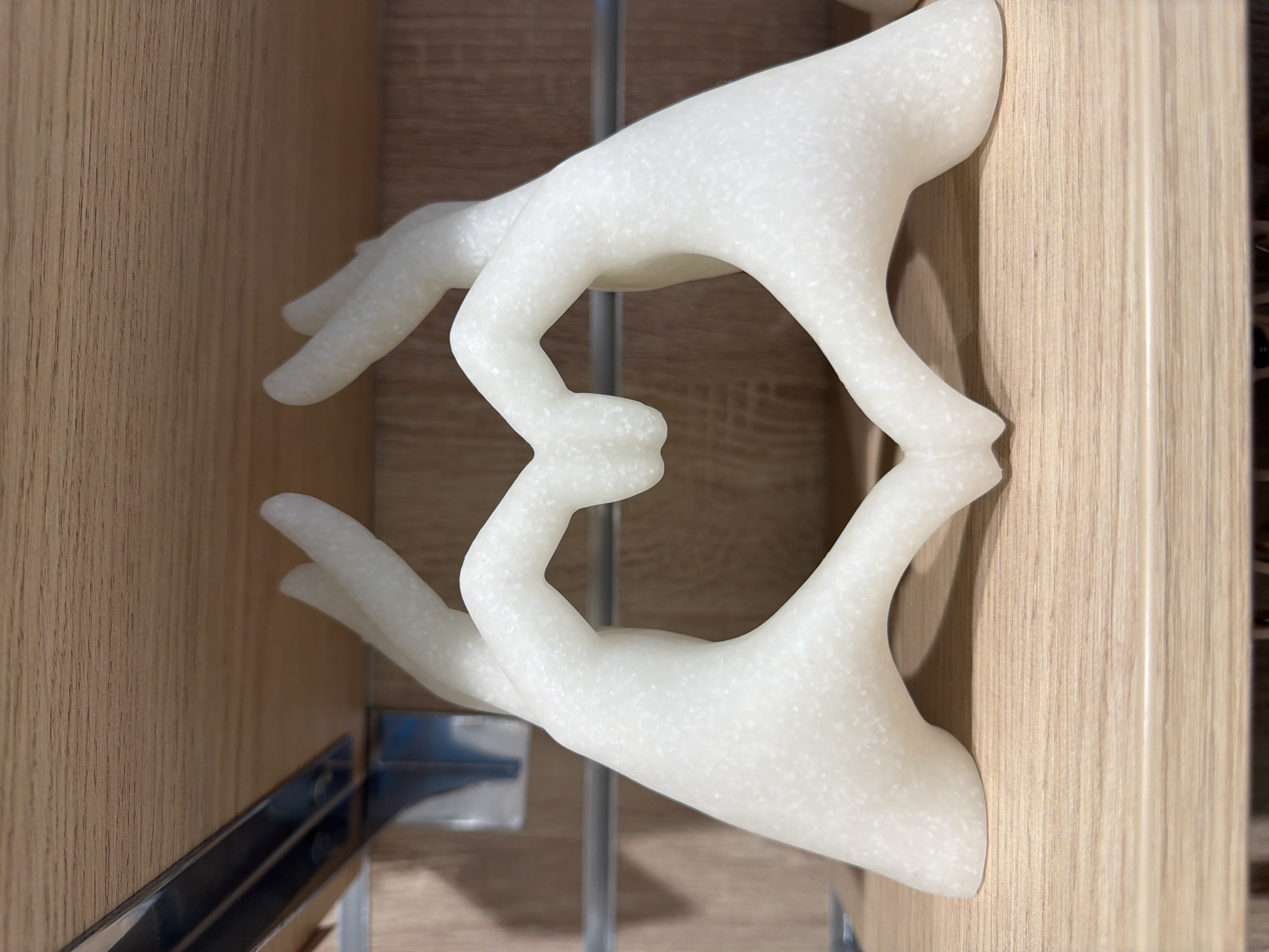 A close-up shot features a pair of white, marble-like hands forming a heart shape with their fingers. The hands are mounted between two vertical, light brown wood panels, likely part of furniture. A metal bracket is visible behind the bottom hand. The location is unknown.