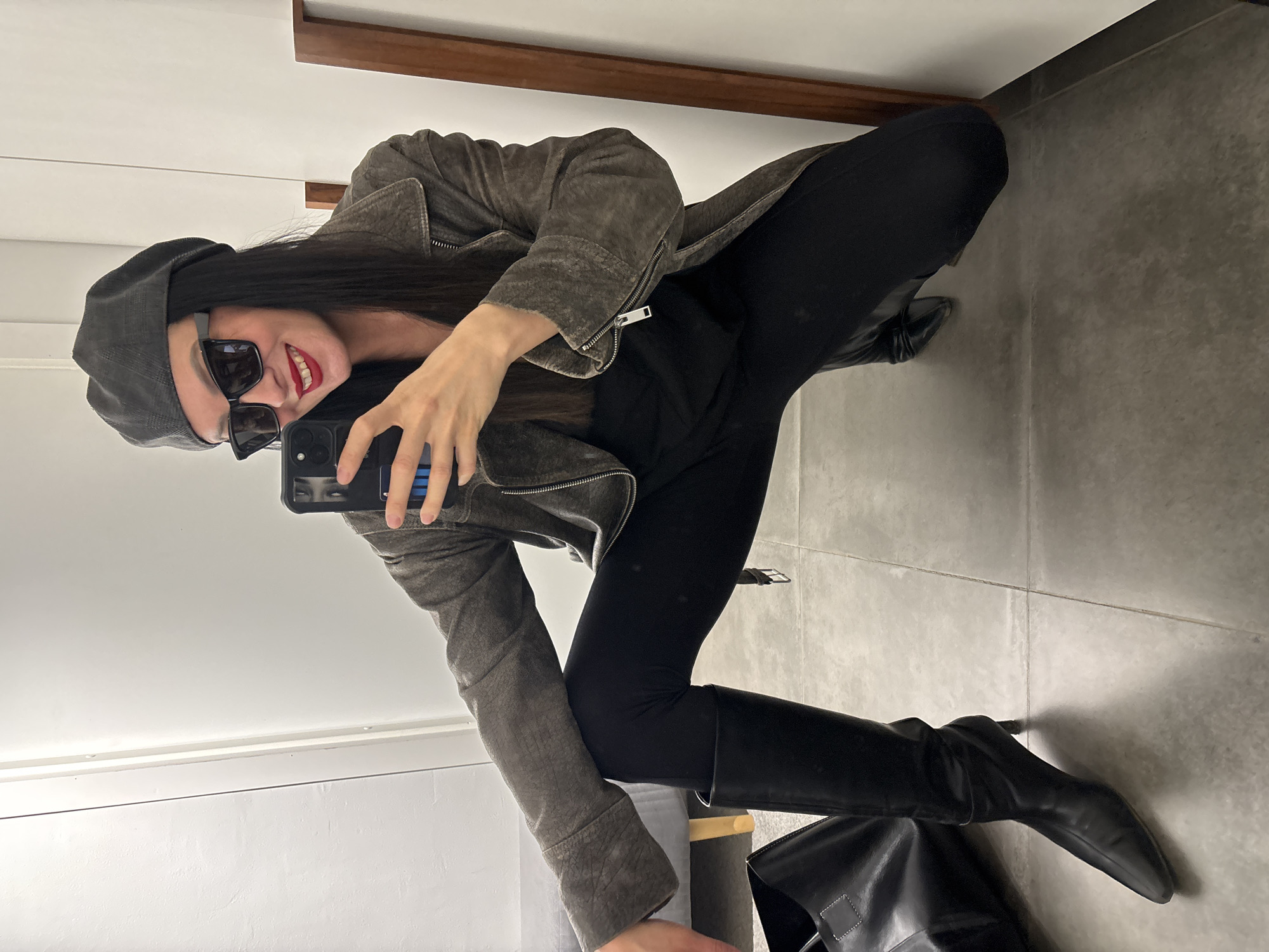 The image shows a woman taking a selfie in what appears to be a fitting room. She is crouching, facing the mirror, holding her phone up to capture the image. She is wearing a grey cap, sunglasses, a grey jacket, black top, black pants and black knee-high boots. In the background, there are walls and floor with tiling. There is also a door in the background. The location is not identifiable as being in Chaikovsky, Russia from the image alone.