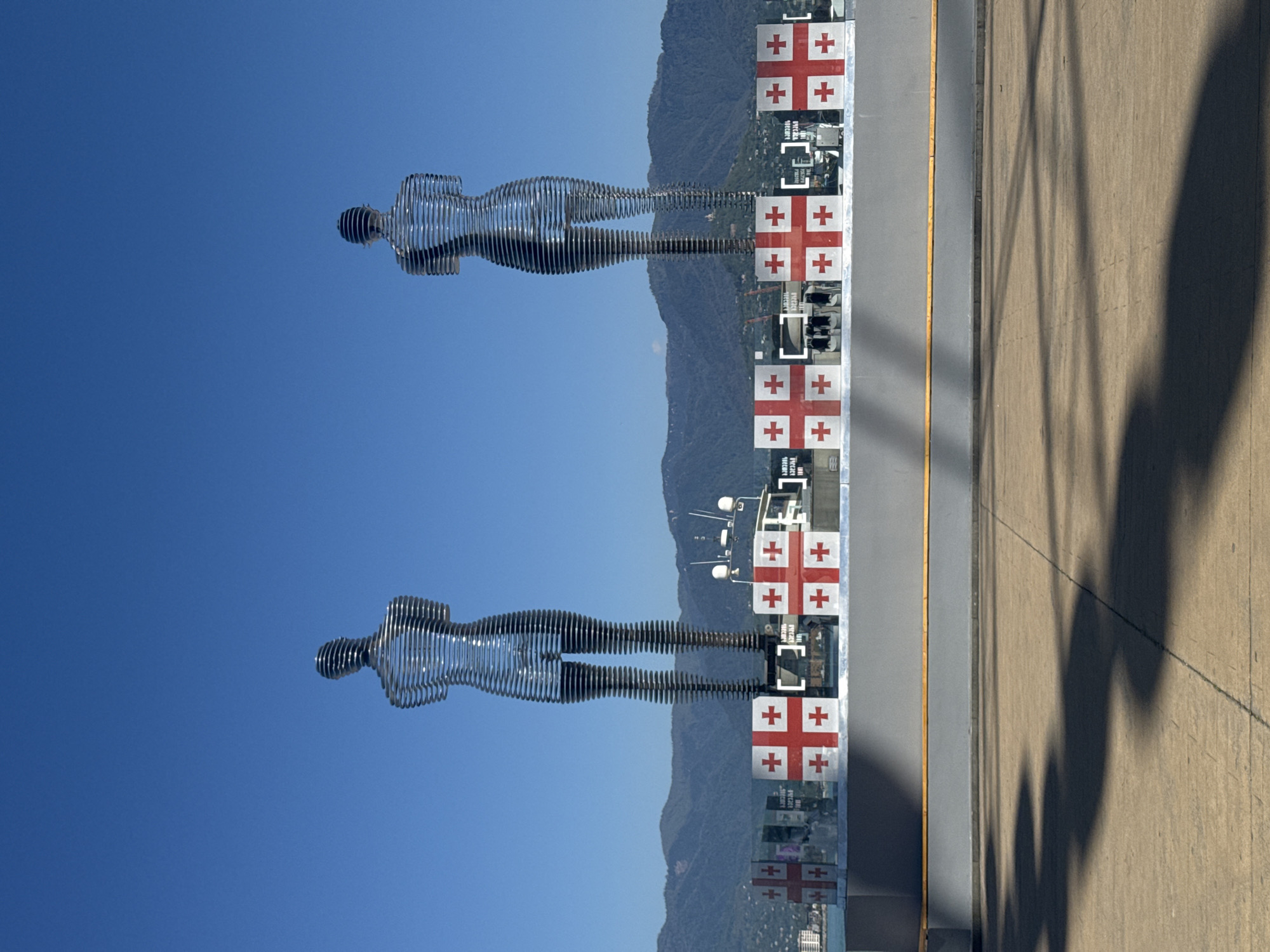 The image depicts the Ali and Nino statue in Batumi, Georgia. The statues, crafted from metal strips, represent a man and a woman. These statues are designed to move and merge into each other. The backdrop includes a clear blue sky and a mountain range. Along a barrier decorated with Georgian flags, shadows of trees are cast on the walkway. The flags have white backgrounds with the red cross design of the Georgian flag.