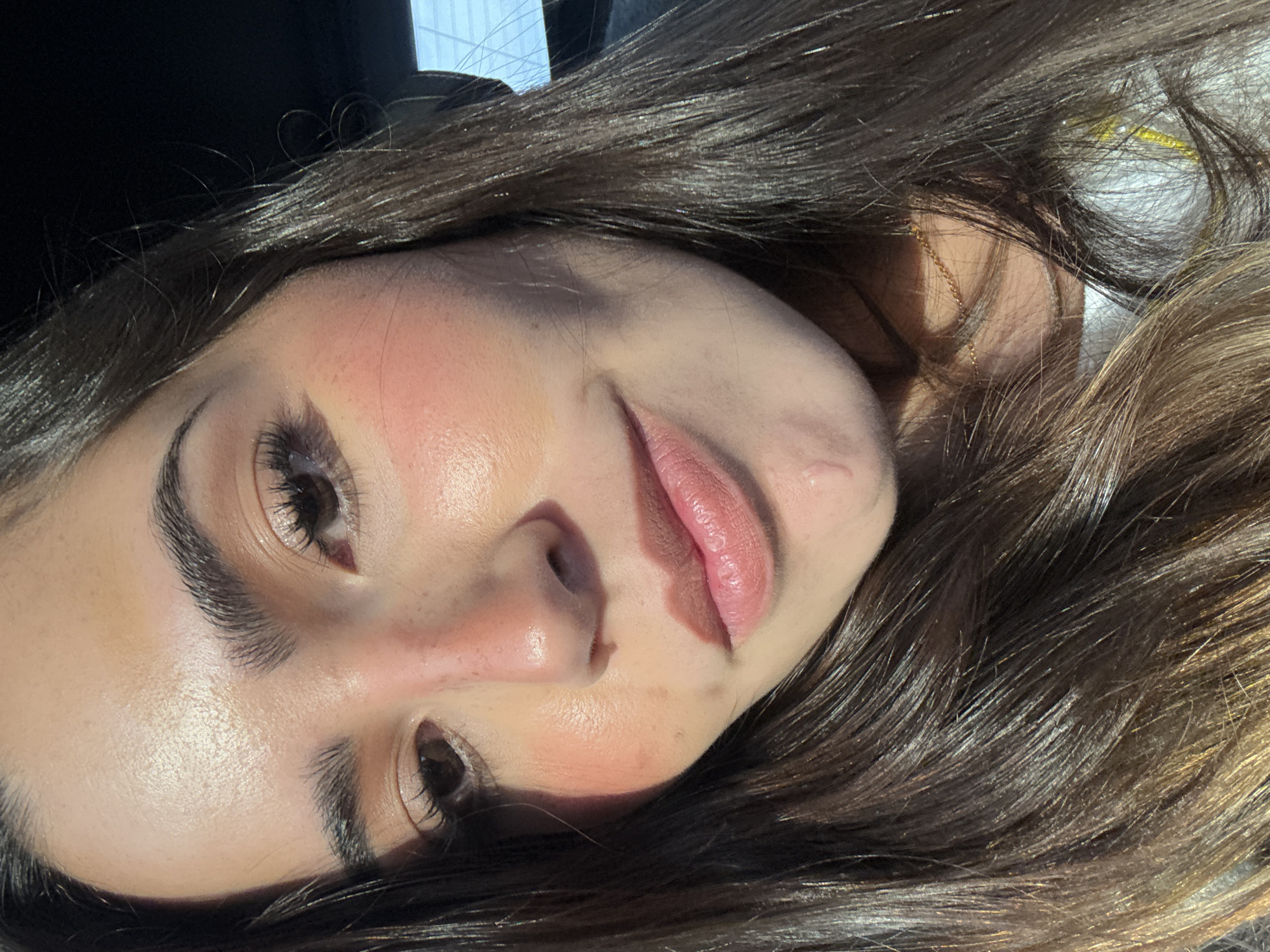 A close-up, angled shot of a woman's face. She has long brown hair, full eyebrows, and appears to be wearing makeup. The lighting is bright. A gold necklace is visible. The location is not discernible from the image.