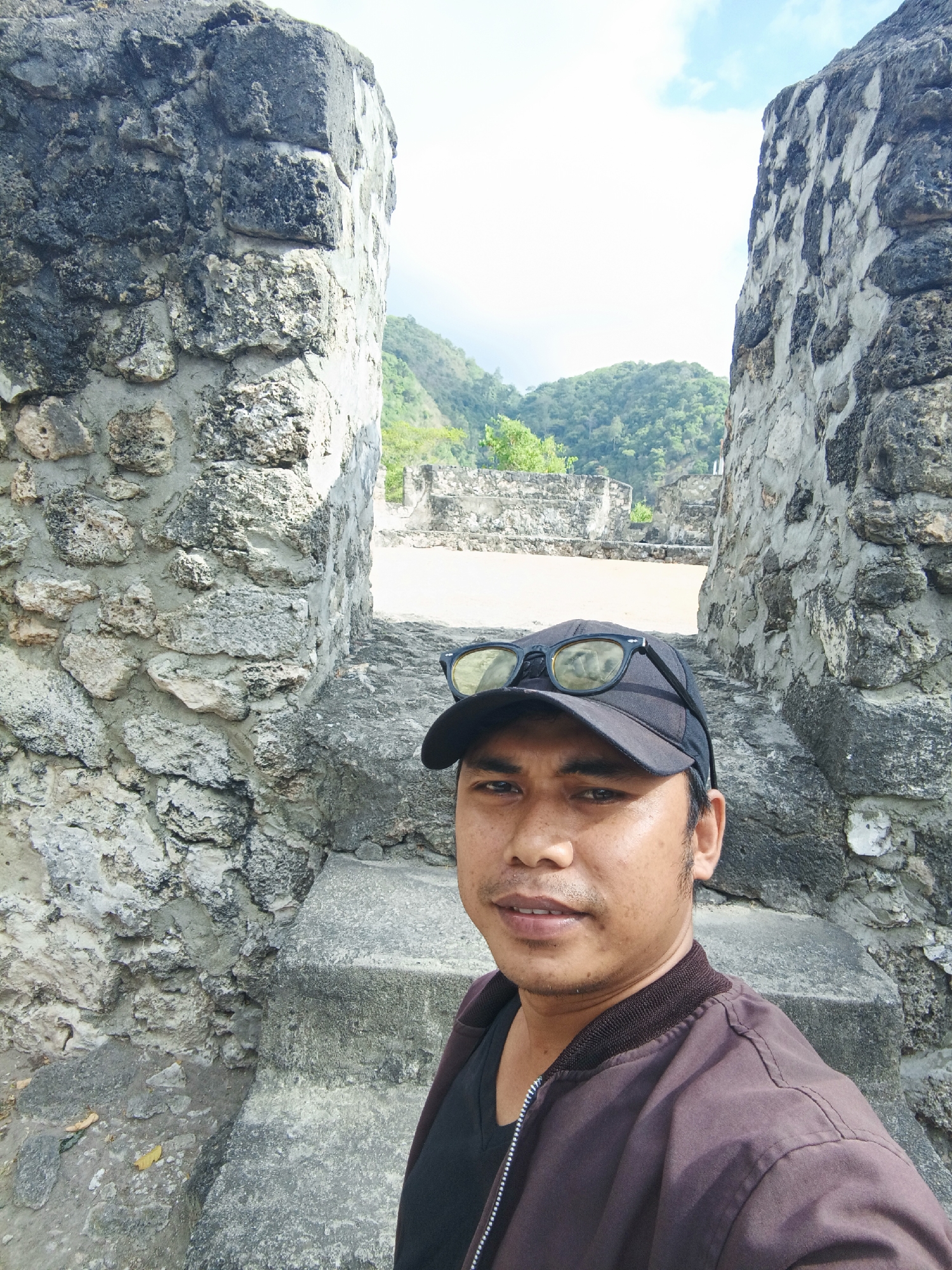 In the city of Gorontalo, Indonesia, a man stands before ancient stone ruins under a bright, partly cloudy sky. He is wearing a black baseball cap with sunglasses resting on top and a dark brown jacket over a black t-shirt. The stone structures, composed of large, rough-hewn rocks cemented together, appear weathered and ancient, with some sections crumbling. In the background, lush green hills and trees rise against the sky, suggesting a natural, perhaps tropical, landscape. The overall scene captures a moment of exploration or sightseeing amidst historical remnants.