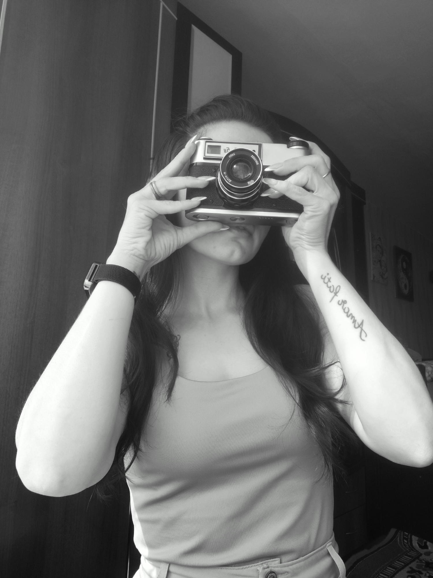 A woman is holding a camera up to her face in a black and white photograph. Her eyes are visible above the camera. She has long dark hair and is wearing a tank top. A black watch band is visible on her left wrist, and a tattoo is on her right forearm. Rings are on her fingers. The background includes a dark cabinet and wall. Based on the provided location, the photograph was taken in Фоки, Russia.