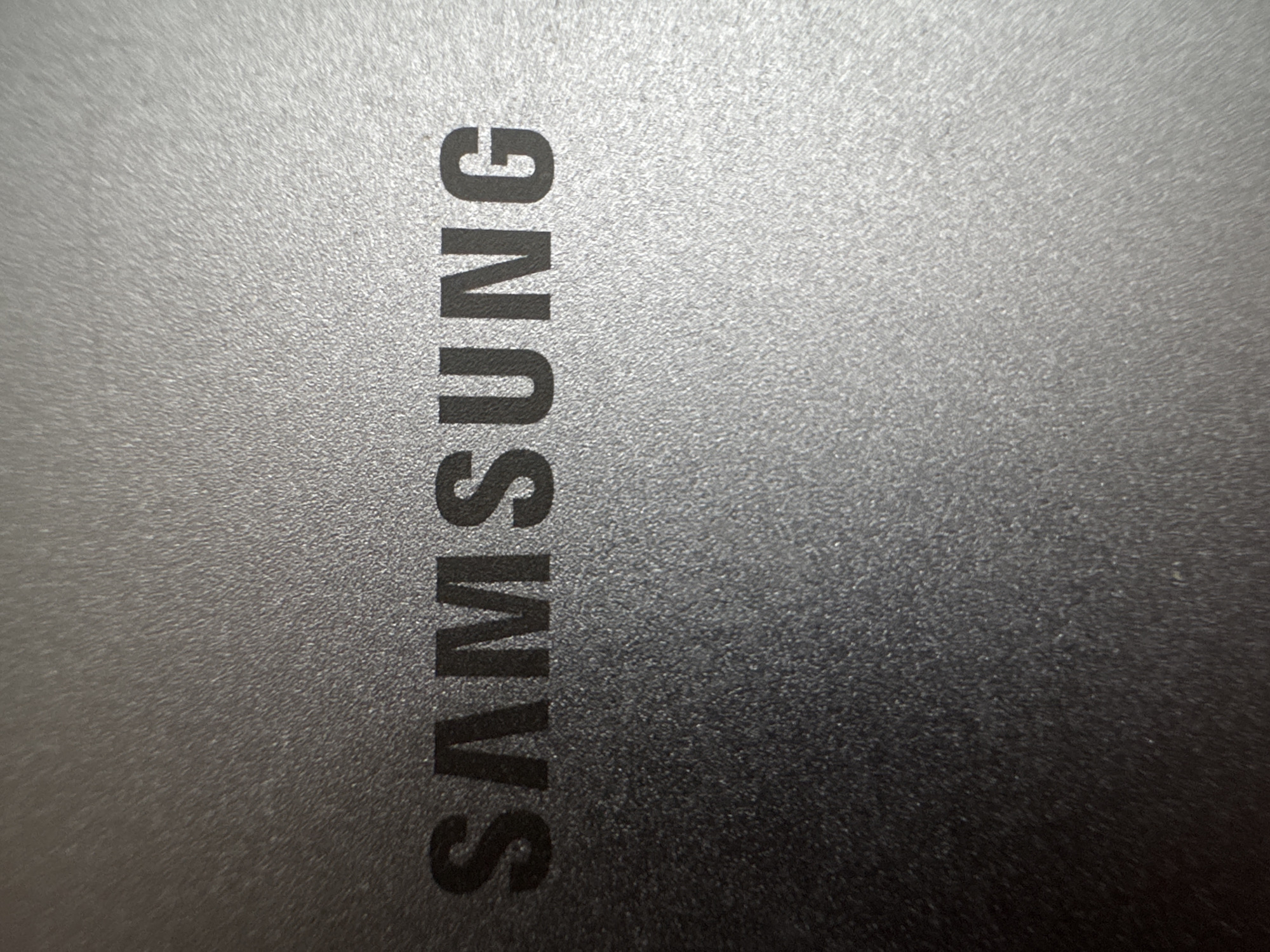 The image is a close-up, vertically oriented shot of the Samsung logo embossed on a textured, dark gray surface. The logo, written in all capital letters, reads "SAMSUNG" and is positioned on the left side of the frame, running from bottom to top. The texture of the surface appears to be a brushed or matte finish, with subtle linear patterns and fine graining, creating a slightly reflective quality that varies with the light. The lighting creates a gradient across the surface, with the left side appearing brighter due to the embossed logo catching more light, while the right side is darker and less detailed. There are no people, specific scenes, or weather conditions visible. The image focuses solely on the product branding. The visible text is "SAMSUNG".