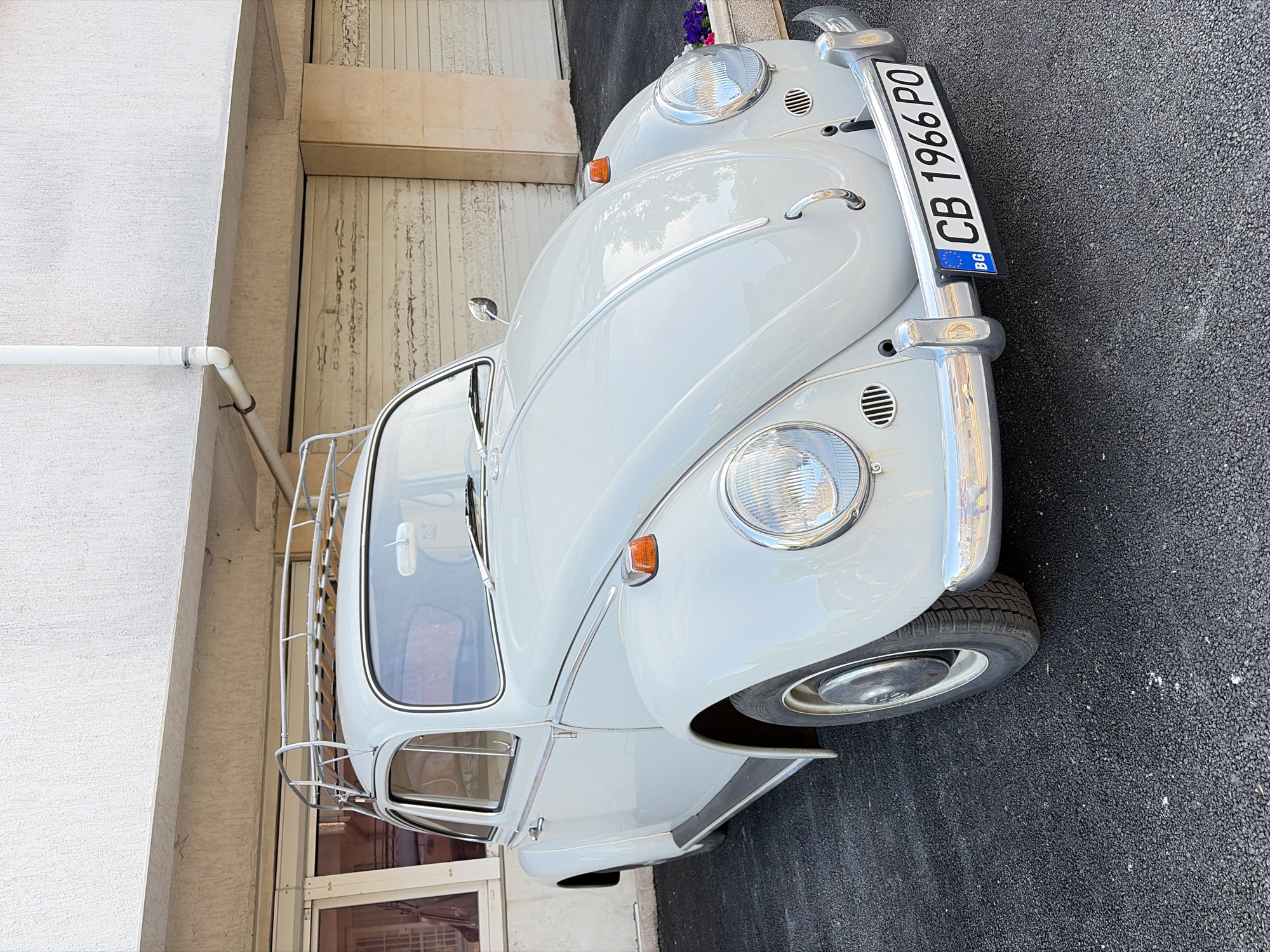 A light gray, vintage Volkswagen Beetle with a roof rack is parked on a dark asphalt surface in front of a building with a beige garage door. The car has chrome accents, including the bumpers, hubcaps, and trim. A license plate reading "CB 1966 PO" is affixed to the front bumper. The building appears to be a residential or commercial property, with a window visible on the left side and a white drainpipe running down the exterior wall. Small planters with purple and pink flowers are situated to the right of the car. The sky is not visible in the frame. The scene is likely in the city of Sofia, Bulgaria, as indicated by the license plate format and the architectural style of the building.