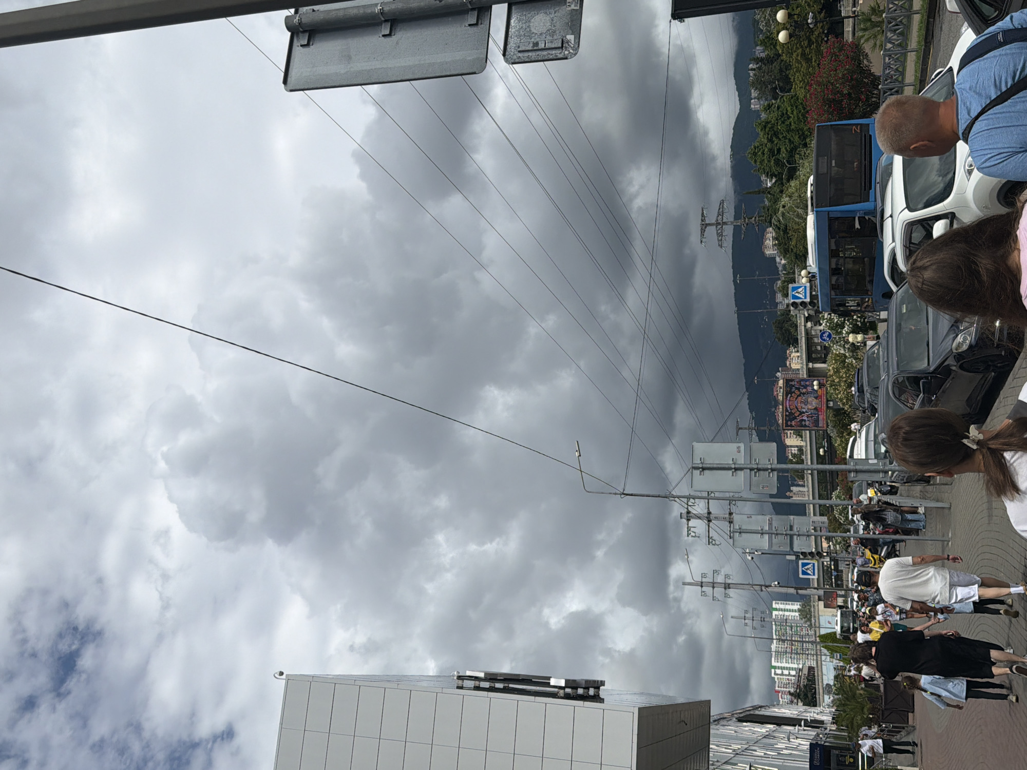 This is a street scene in Sochi, Russia, on a cloudy day. A modern building with white tiled facade is on the left. A group of people are walking on a paved walkway in front of the building, with some people walking away from the camera and others walking towards it. There are cars and a blue bus on the street. In the background, there are buildings and lush green mountains under a dramatic sky filled with dark, heavy clouds.